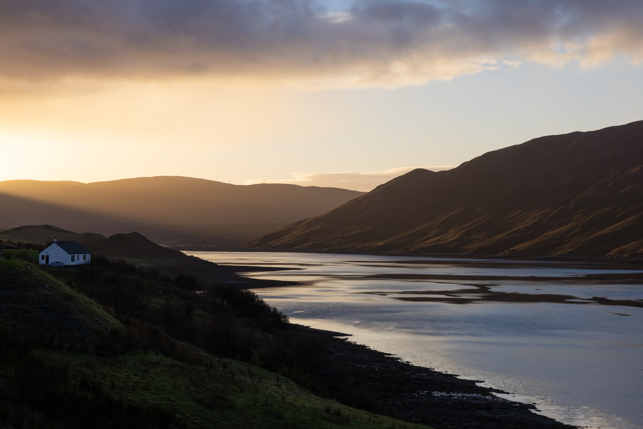 Golden sunset light over a calm estuary with winding sandbanks, dark mountain ridges in the distance, and a small white house on a grassy hillside in the foreground