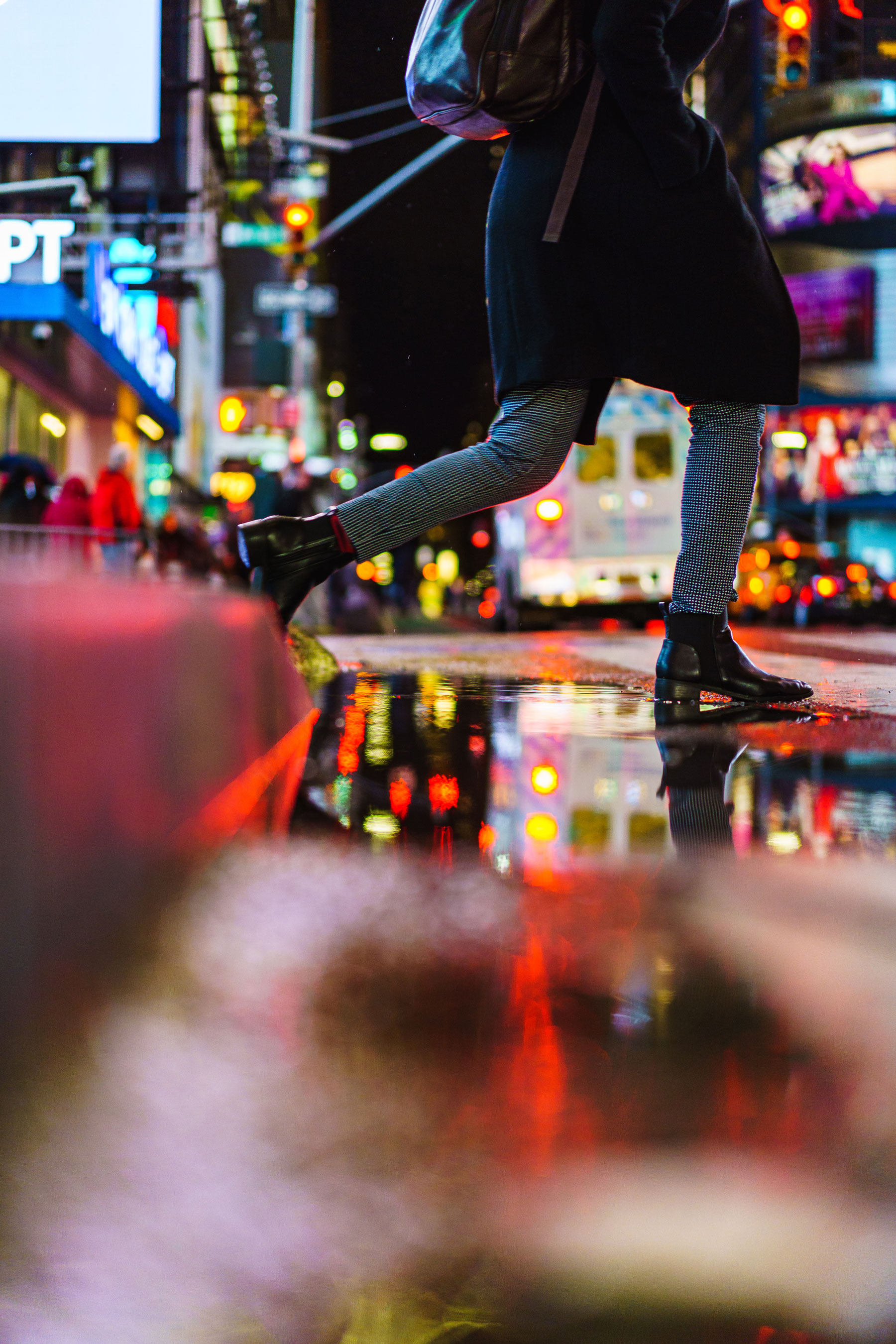 Person stepping over puddle surrounded by bright city lights at night, photo by Manuel Pena on a Sony A7 III.