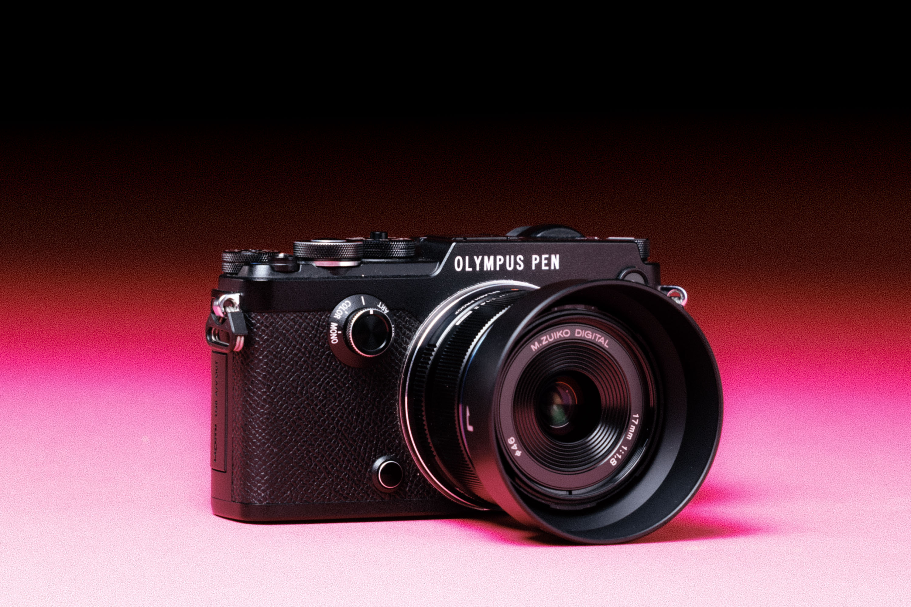 A used Olympus Pen-F camera with an M.Zuiko 17mm lens, with an added film-like grain effect on a graduated pink background. 