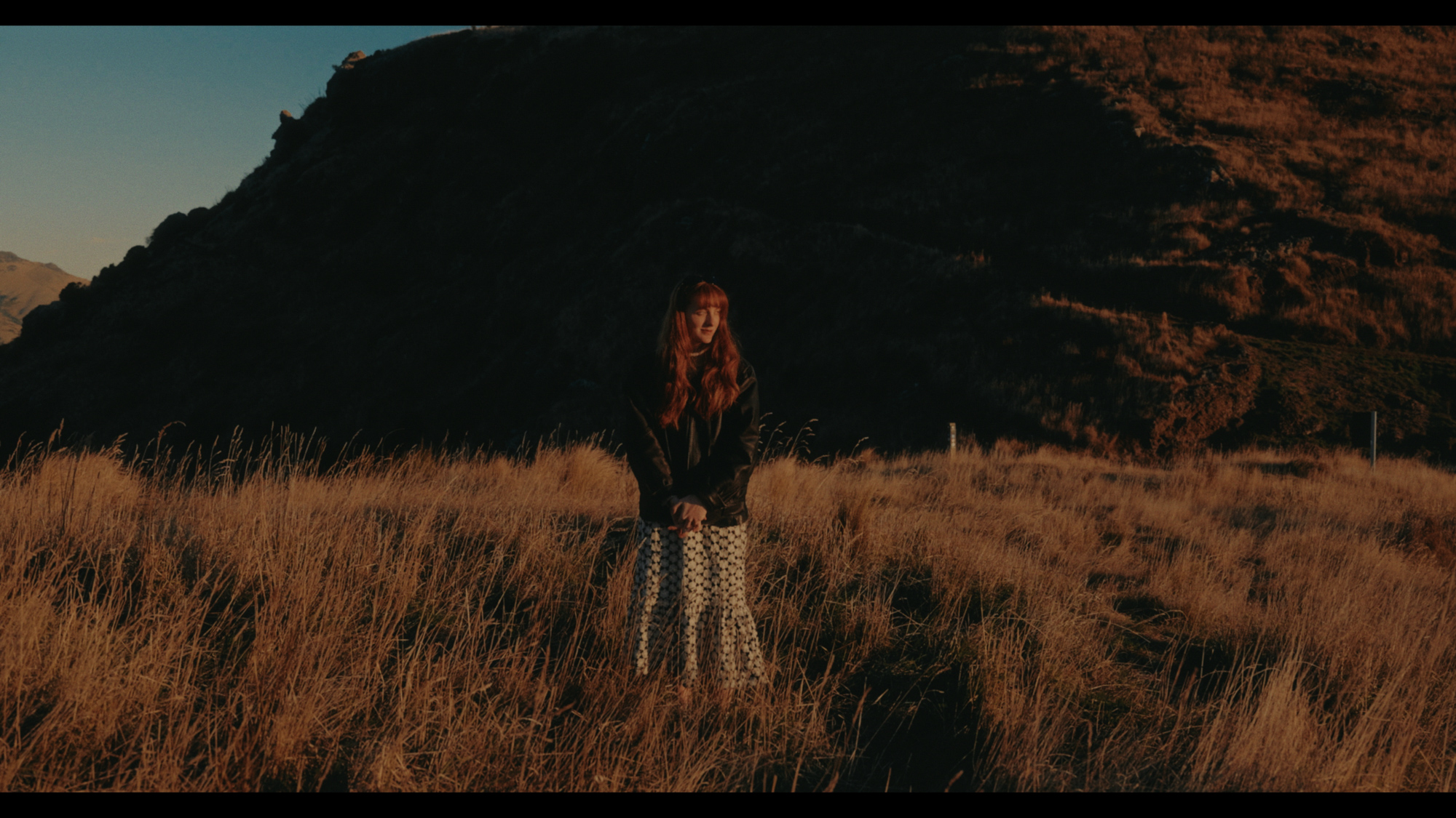 A person with long reddish hair wearing a black jacket and patterned skirt stands in a dry grassy field.