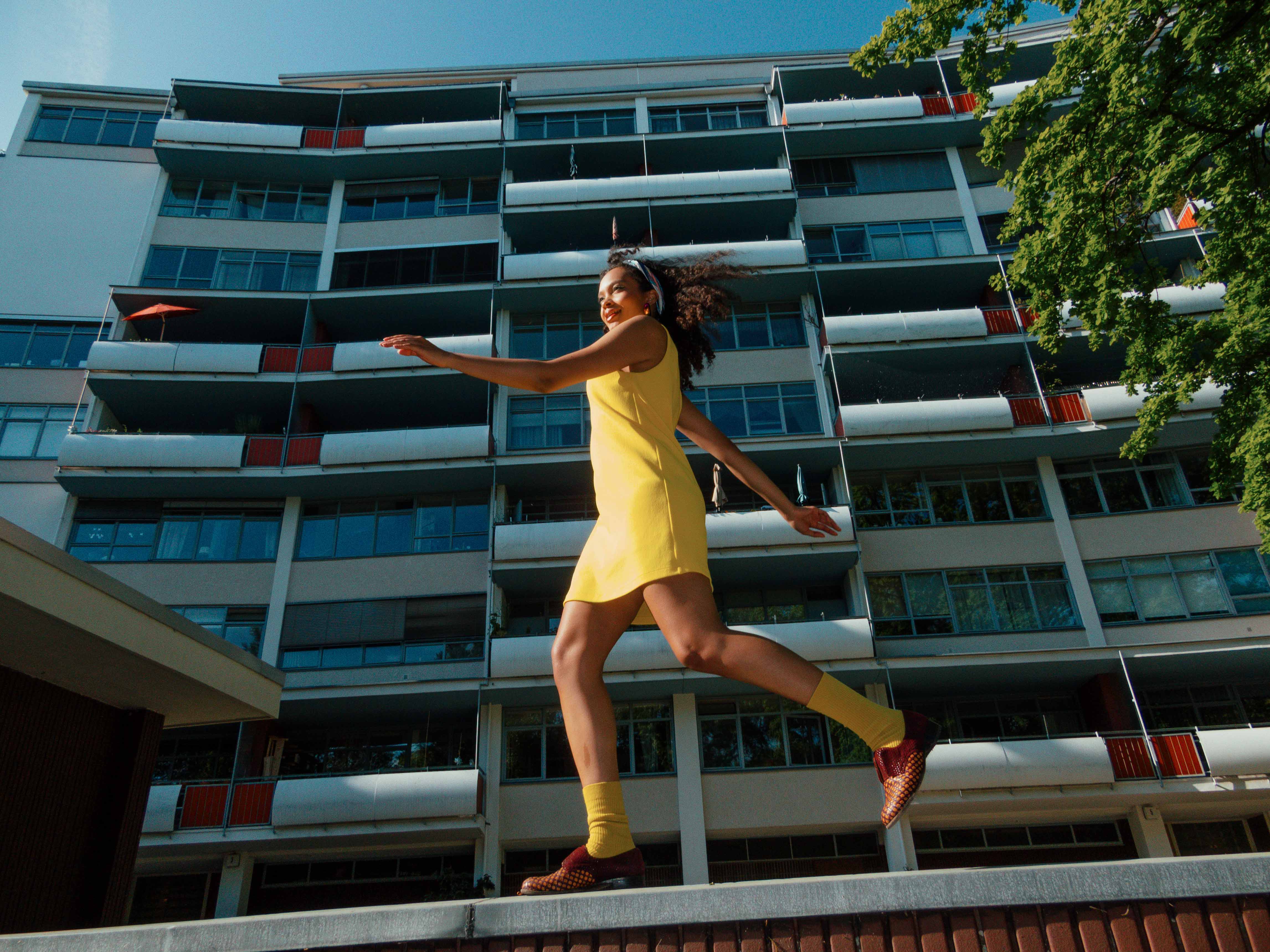A model in a yellow dress and matching socks, playfully walking on a wall in front of a modernist building in Berlin’s Hansaviertel. Shot by Alina Rudya on a Nikon Coolpix A.