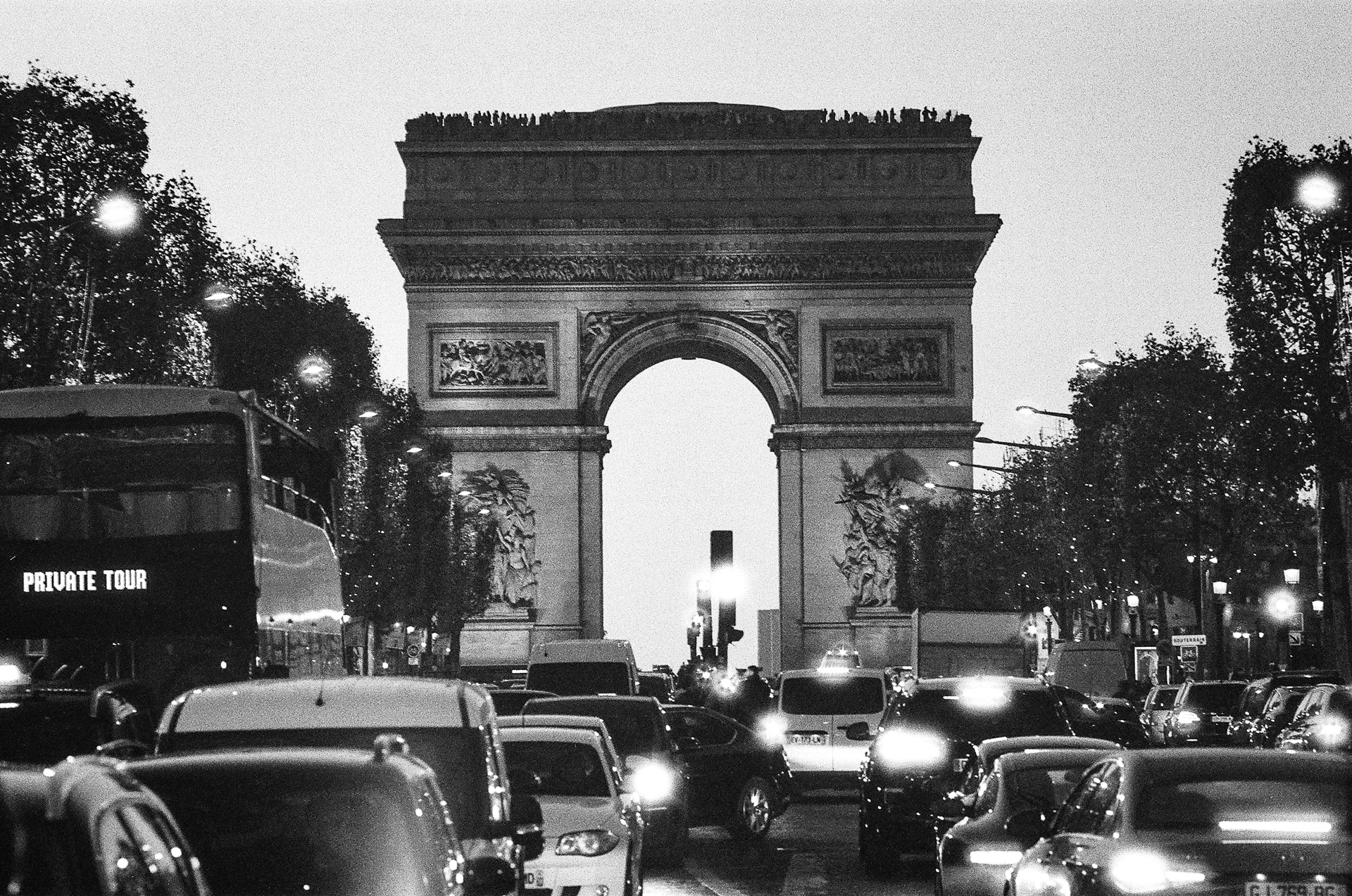 Black and white view of the Arc de Triomphe in Paris with busy traffic, glowing headlights and evening light creating a dramatic urban city scene.