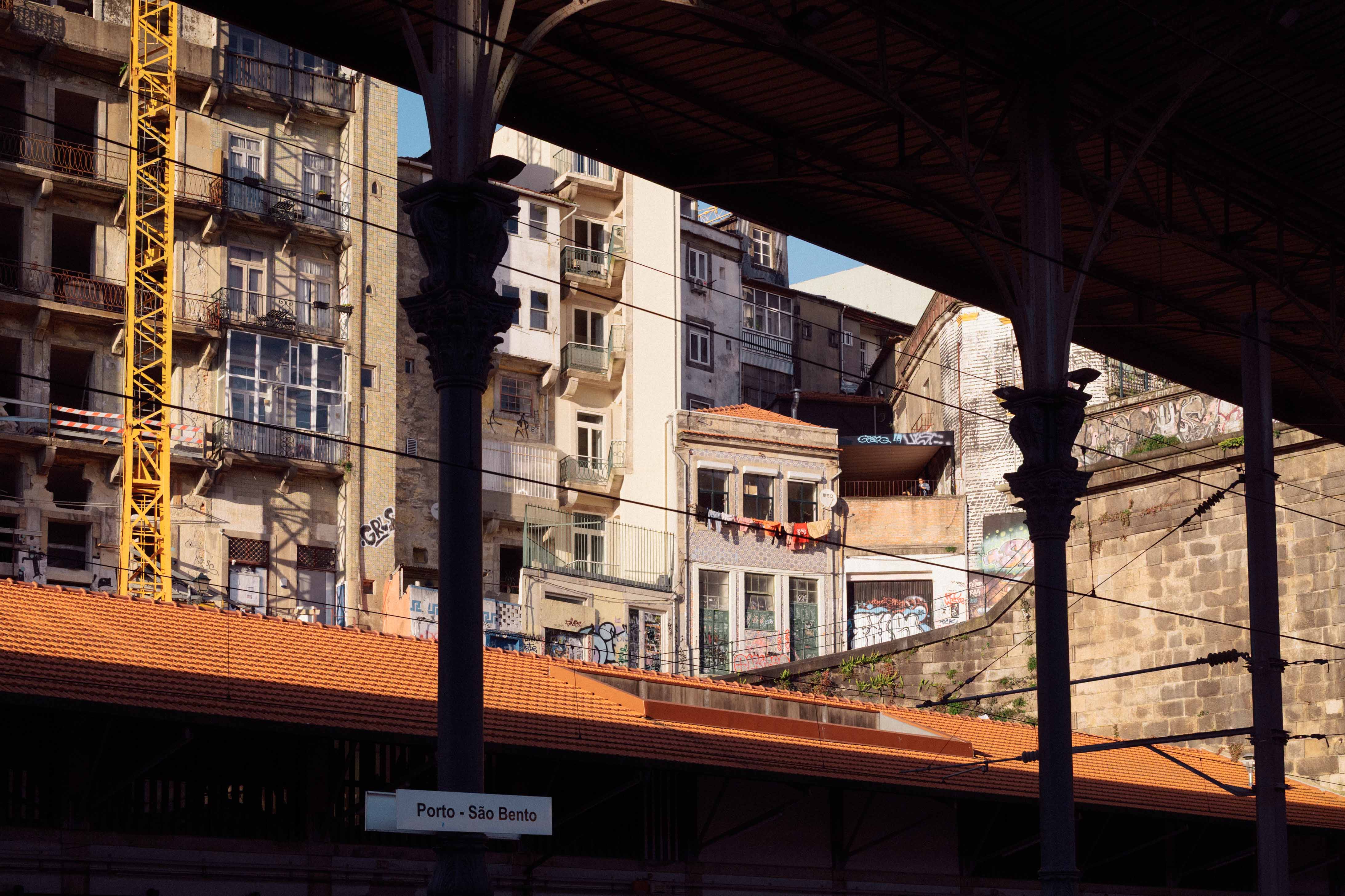 View from São Bento station platforms looking towards weathered residential buildings stacked above the tracks, framed by iron columns and overhead cables.
