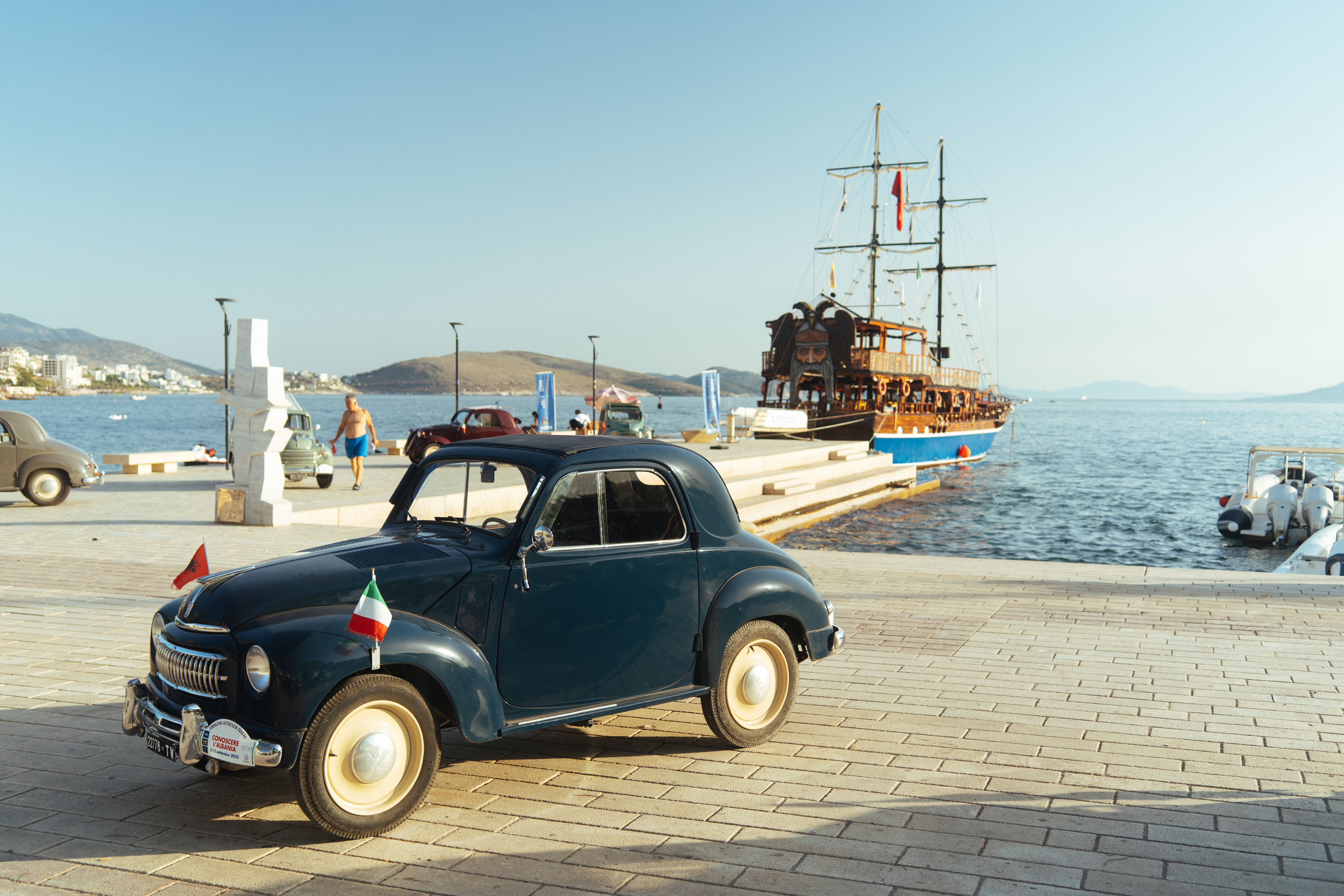 A vintage car sits by the waterfront, with a traditional sailing ship and calm sea in the background.