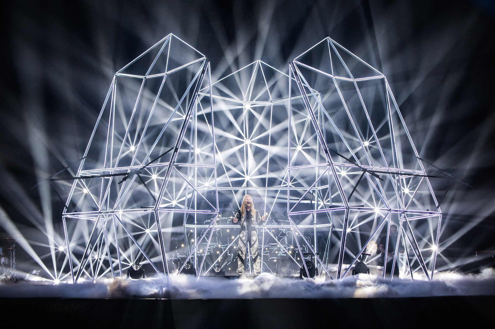 A performer at Eurovision standing under a large metal structure.