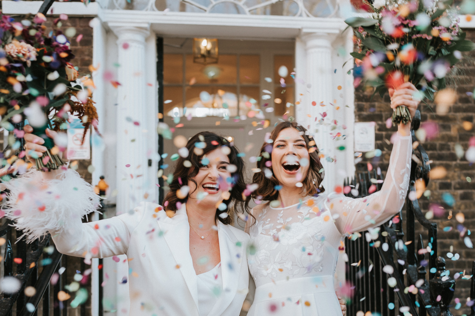 
A wedded couple, one dressed in a white suit with fluffy arm cuffs and one in a white dress, celebrating their wedding day with bouquets of flowers and confetti outside their wedding venue, image by Stephanie Large, Shot on Sony A7 IV

