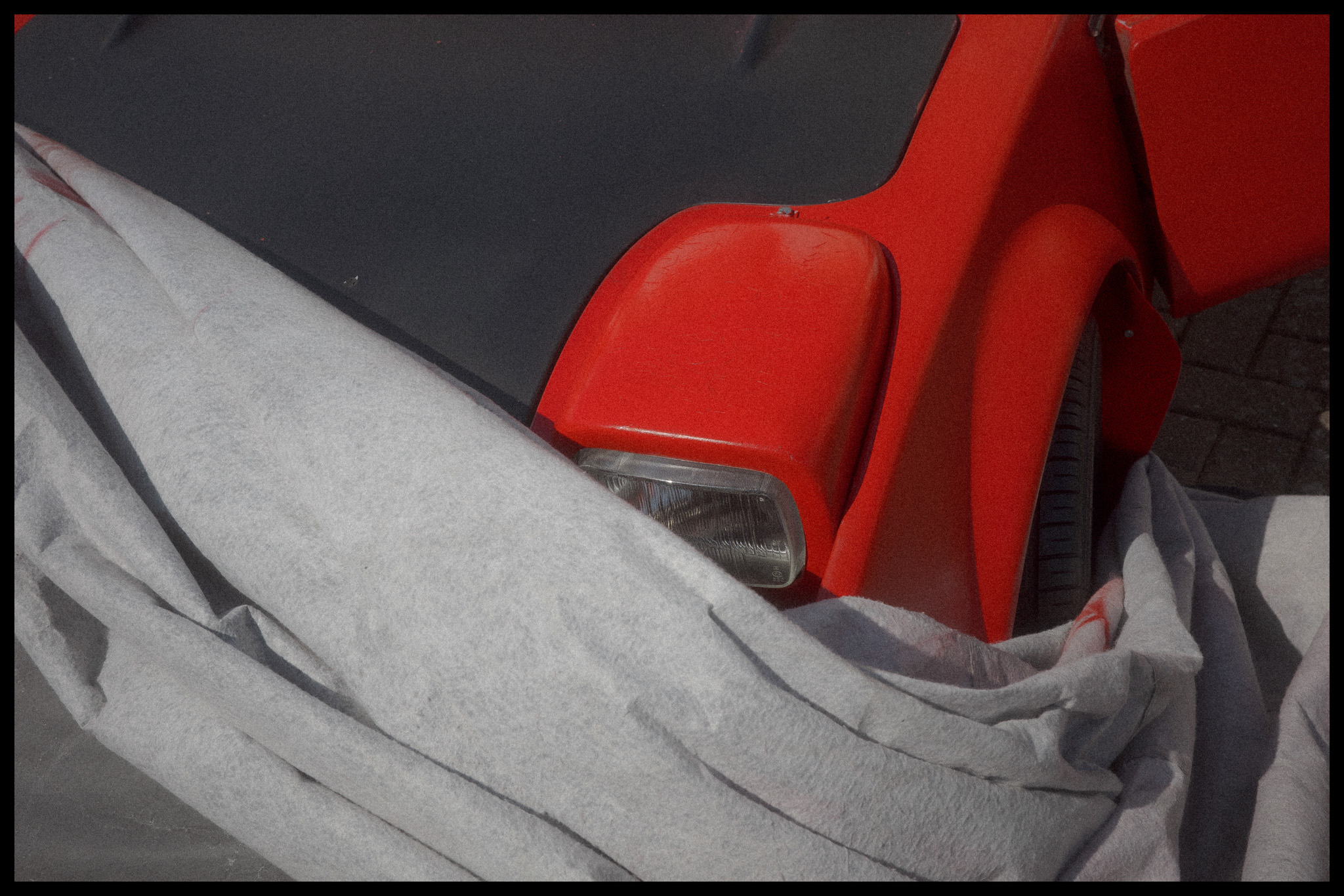 A vintage-looking red car’s headlight with a grey wrap half covering it by Adam Hince