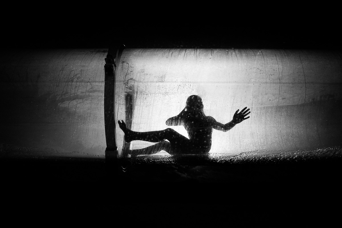 A silhouette of a person inside of a slide raising their hand to the edge of the slide, black-and-white street photography by Marco Larousse