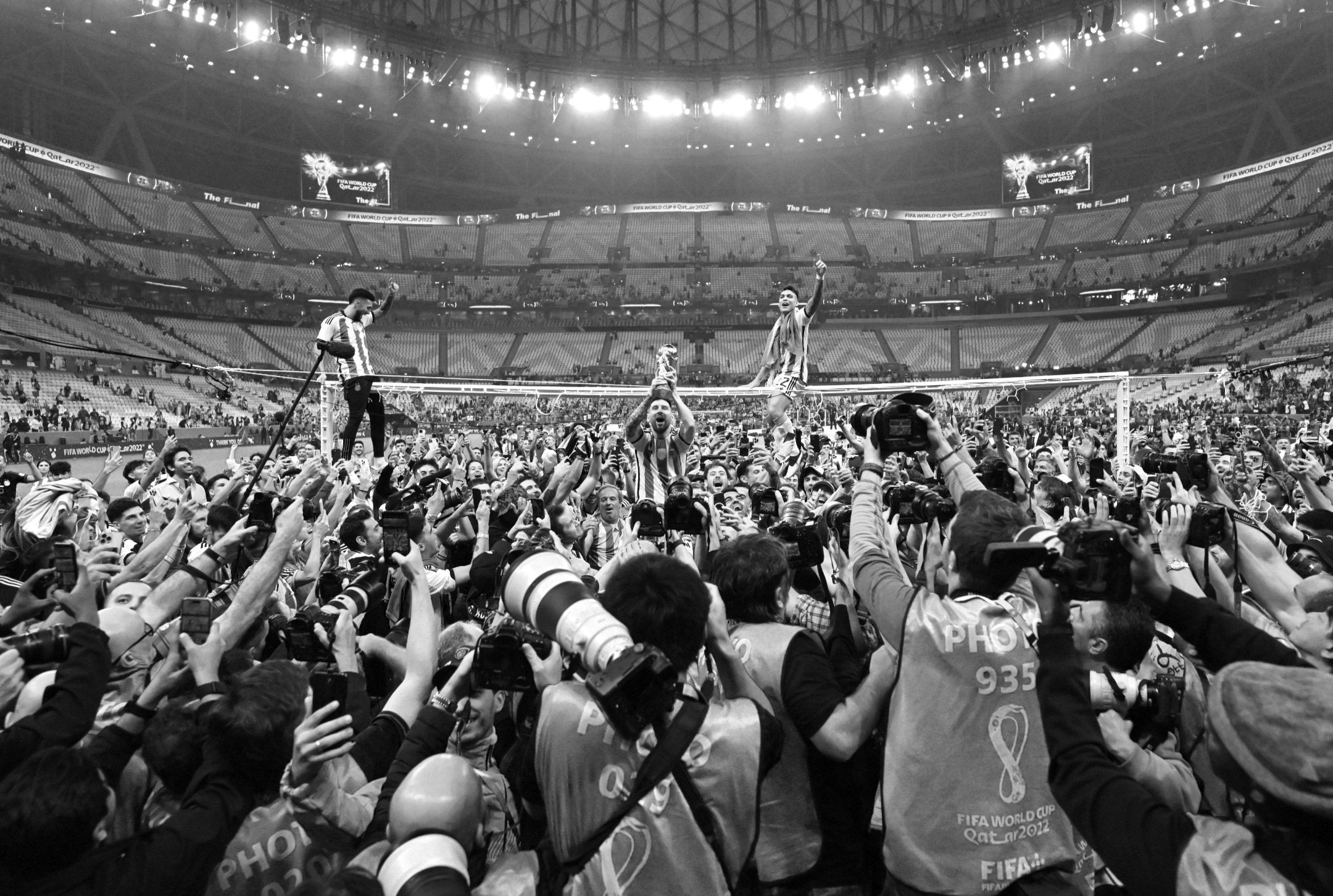 Photo en noir et blanc de Lionel Messi brandissant la Coupe du monde, entouré de ses coéquipiers argentins, de supporters et de photographes, à l’issue de la victoire de l’Argentine contre la France en finale de la Coupe du monde 2022.