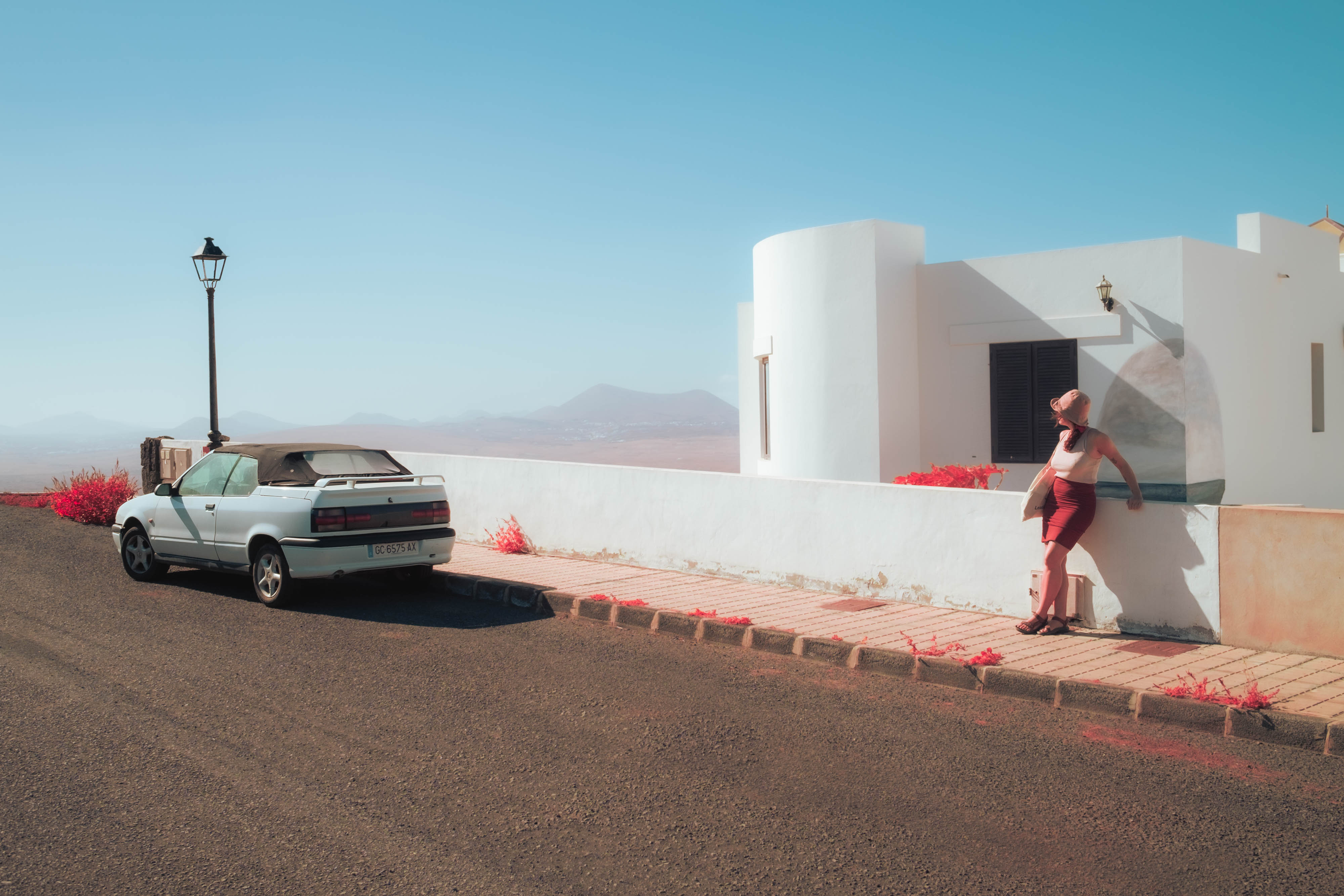 An infrared photo of a car parked on a street next to a white house with someone leaning against a white wall in Lanzarote, shot by Ian Howorth with an IR-Converted Fujifilm X-T1