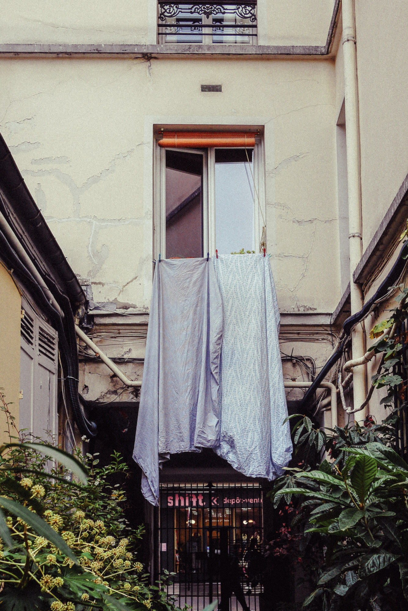 Streetaufnahme von Bettlaken, die umgeben von Wänden, Rohren und Grün aus einem offenen Fenster über einem schmalen Pariser Innenhof hängen. Aufgenommen von Wesley Verhoeve mit einer Panasonic Lumix GF1.