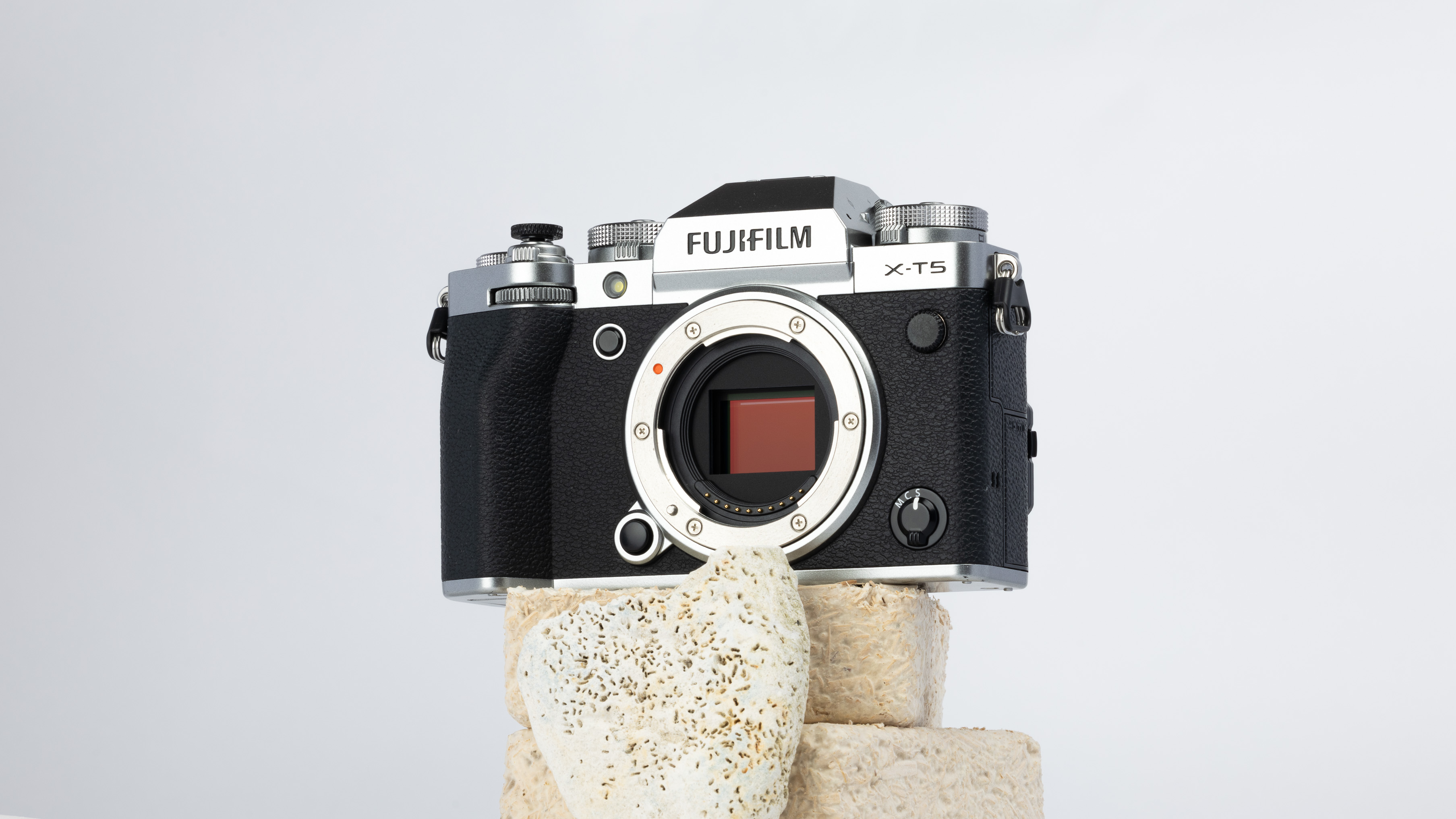 A product shot of a secondhand Fujifilm X-T5 camera propped up on a rocky podium on a white background