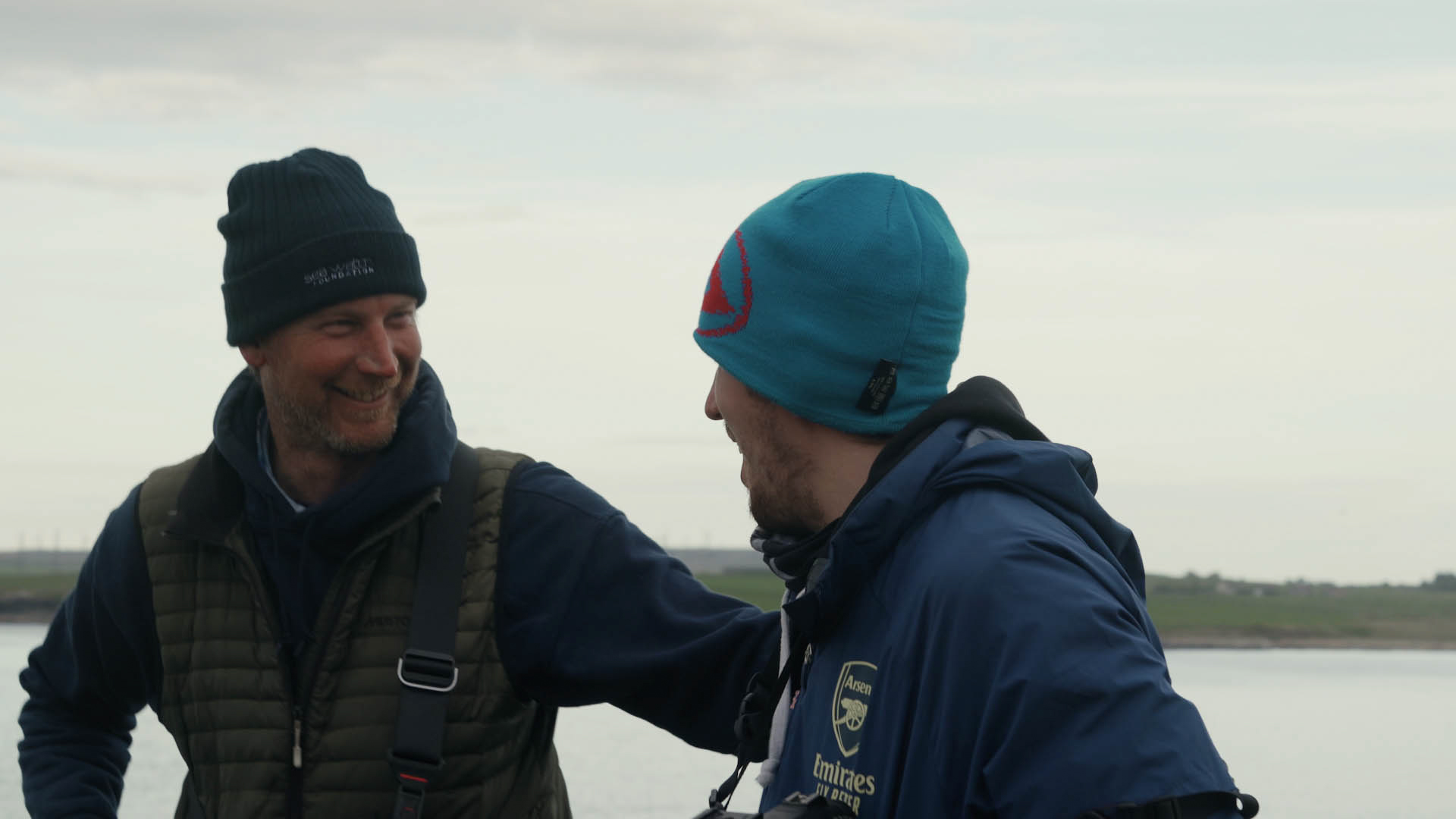 Twee mensen met waterdichte jassen en hoeden omhelzen elkaar bij de zee, een still uit de korte film Truluck van Matt Cannon en Jake Smallwood.