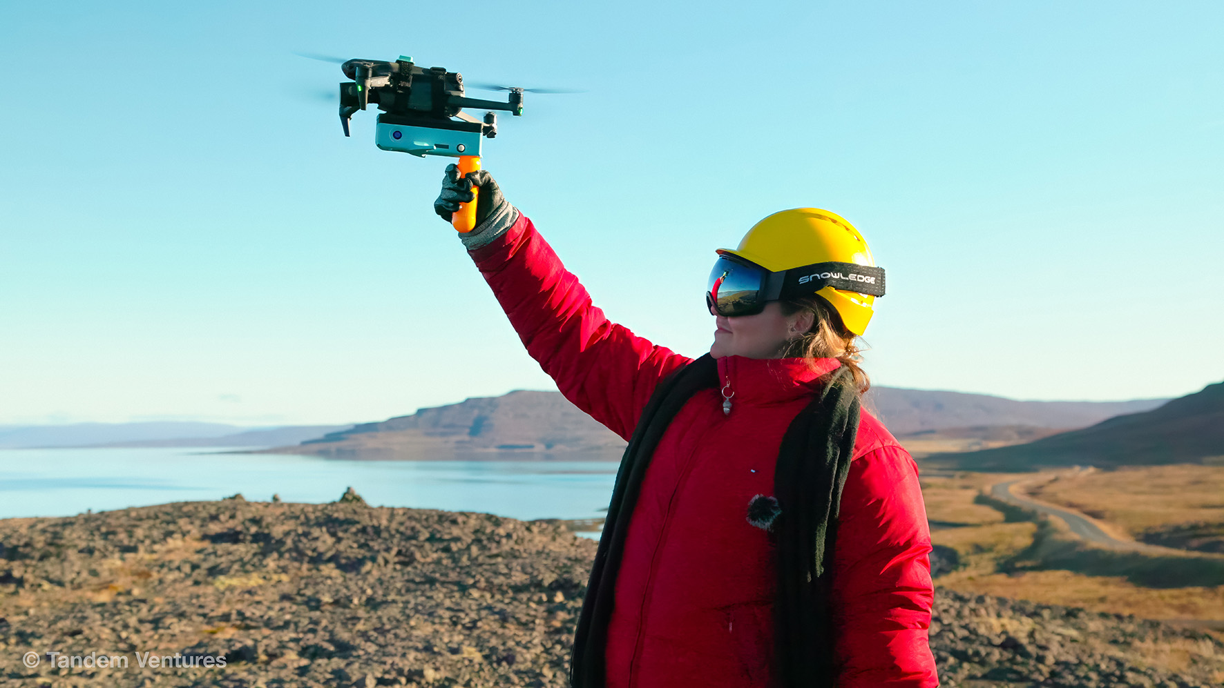 A person wearing a yellow helmet and protective eyewear holds a piece of equipment aloft, surrounded by rocky terrain, blue sky and water.