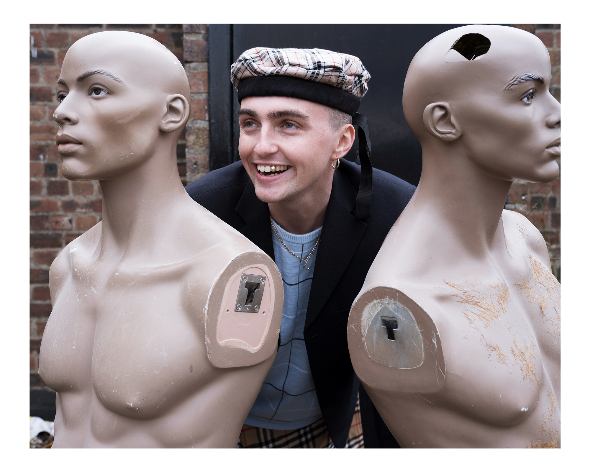 A colour portrait photo of a model wearing a tartan tam o' shanter hat standing between two mannequins in front of a brick wall, photo by Simon Murphy with the Fujifilm GFX 100.
