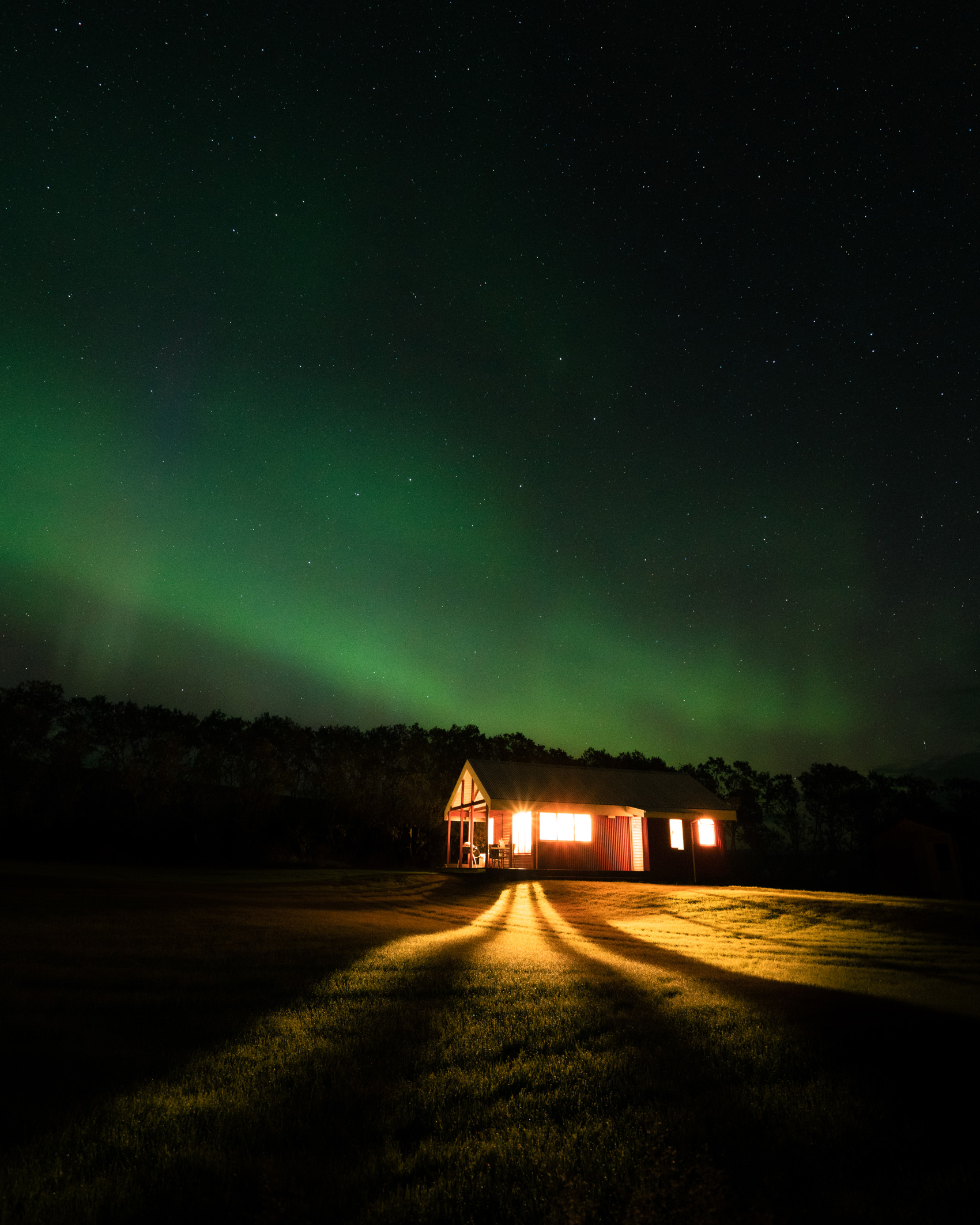 A long exposure of the bright windows of a house in Iceland lighting up the ground at night while the aurora borealis shimmers green overhead, shot on the Sony A7R III by Jordan Lancaster-Graham