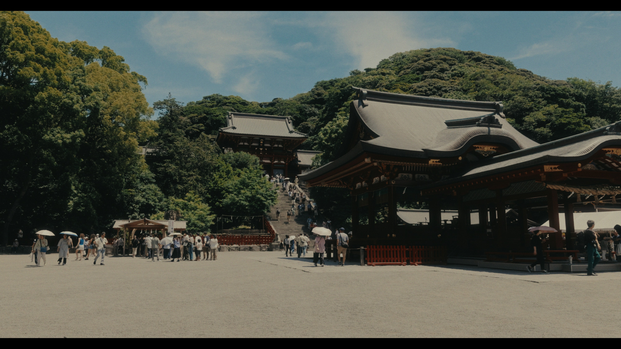 Traditional Japanese temple buildings stand at the base of a forested hill, with visitors gathered on the gravel courtyard.