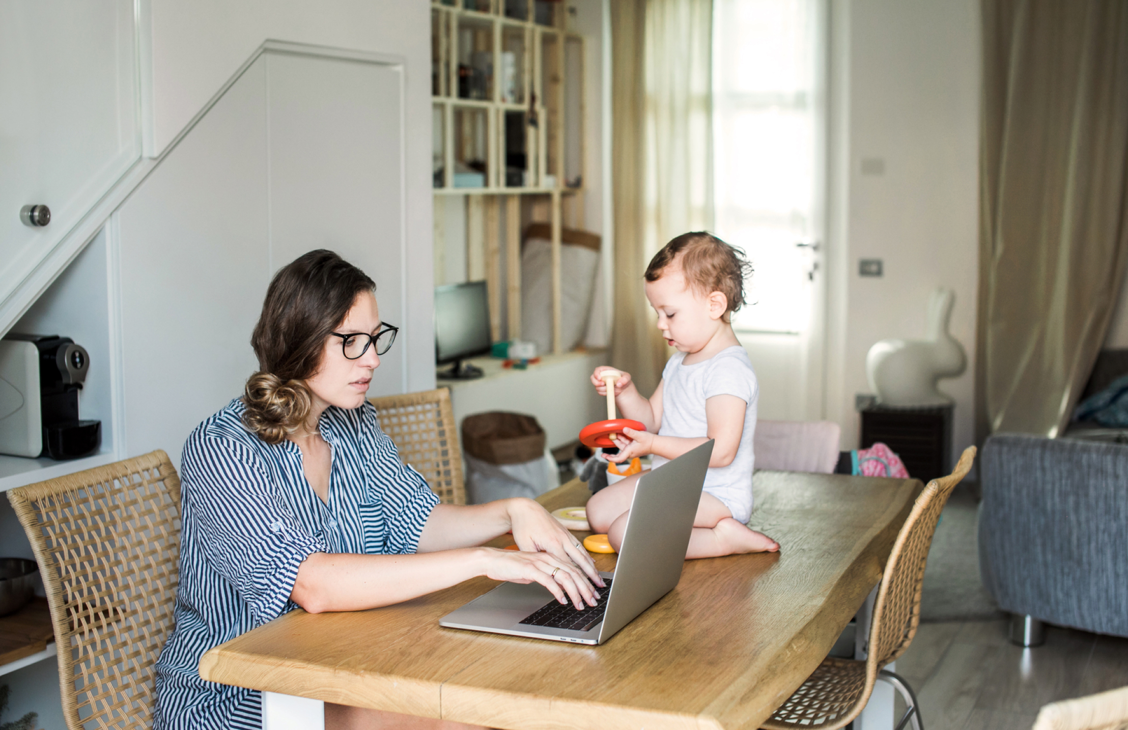 Photo of woman working on her laptop at a table with her baby