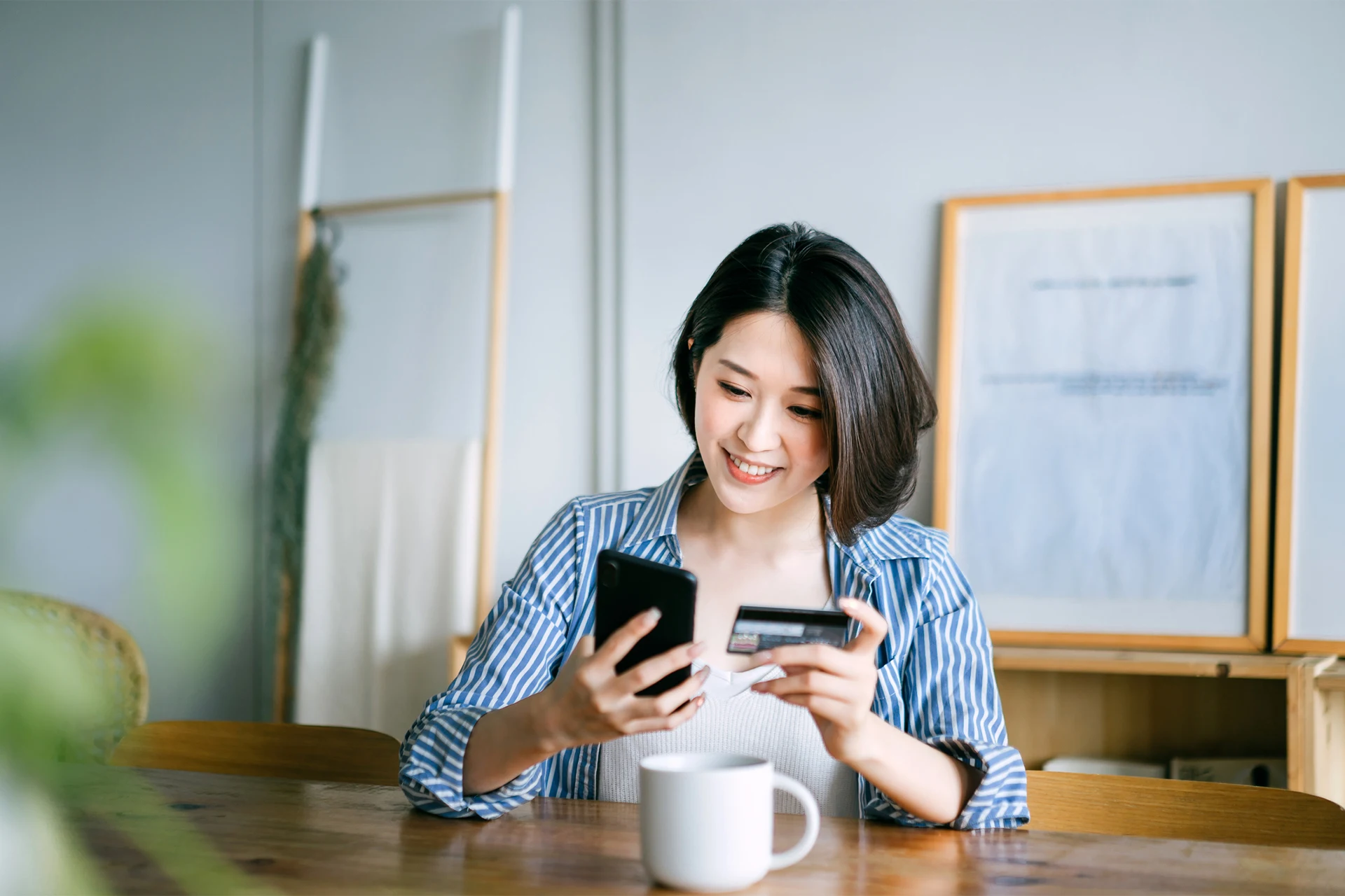 Person holding a smartphone and a credit card while sitting at a table with a mug