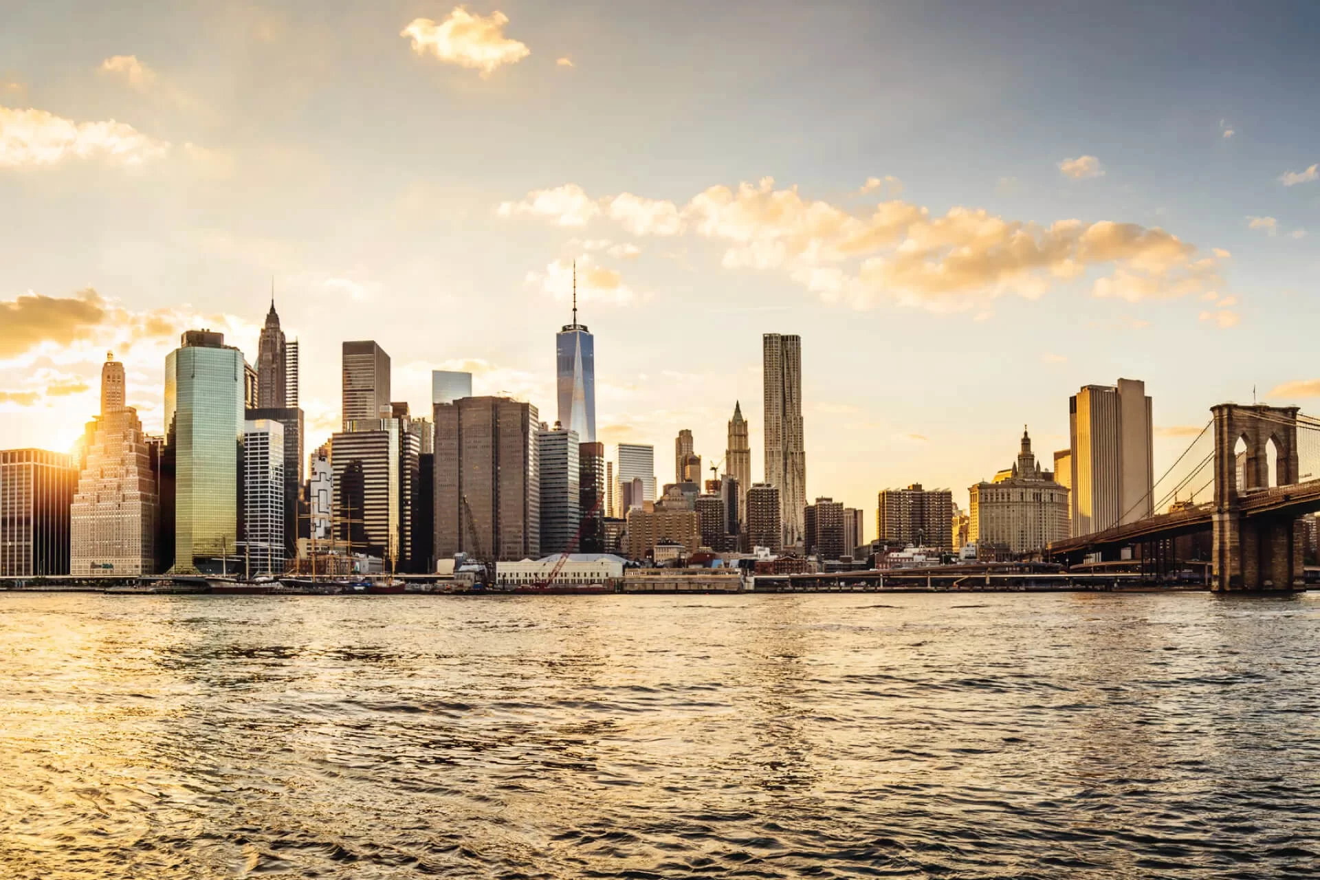 Skyline of a city at sunset with a river and bridge in view.