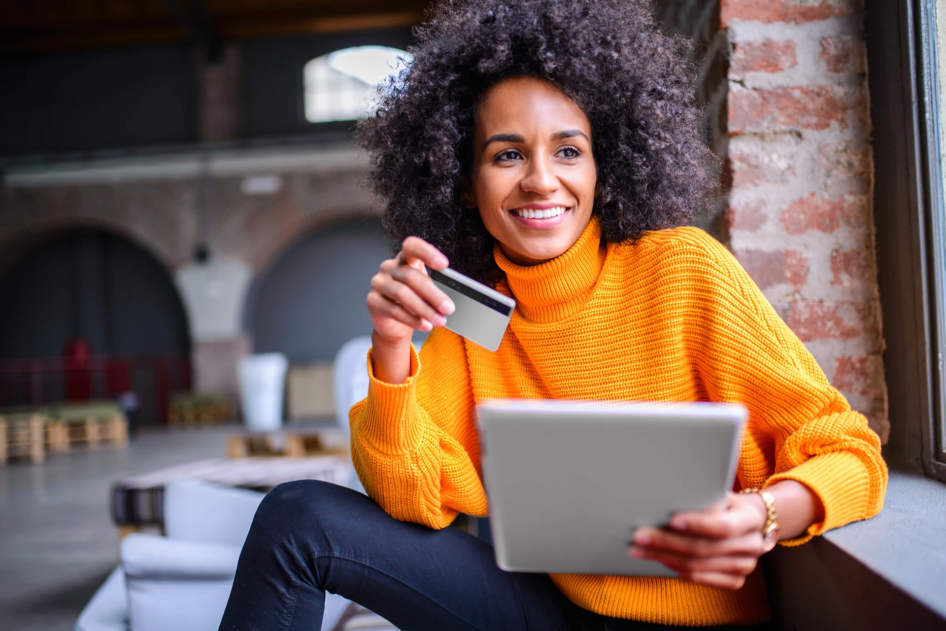 Person holding a credit card and tablet while seated by a window.