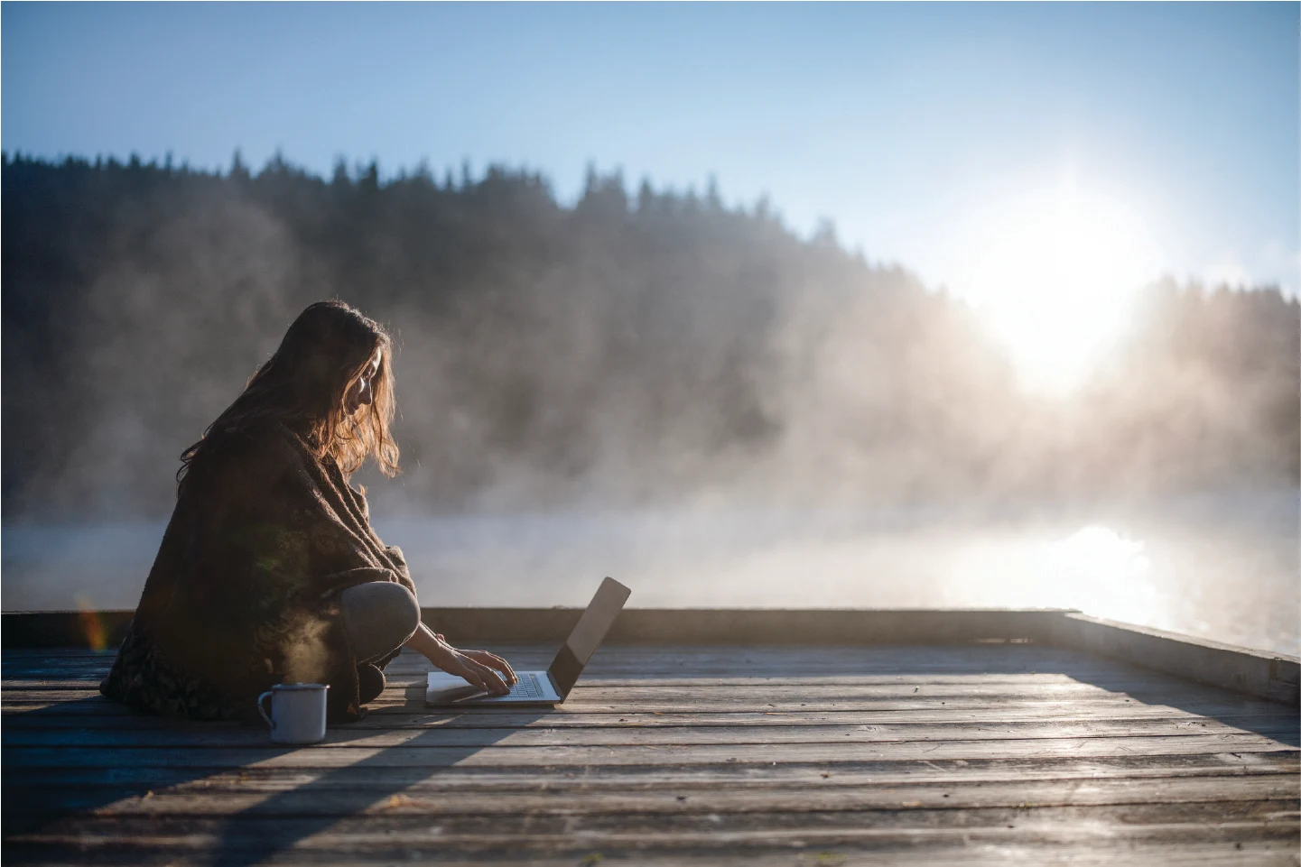 Person using a laptop on a dock by a misty lake at sunrise