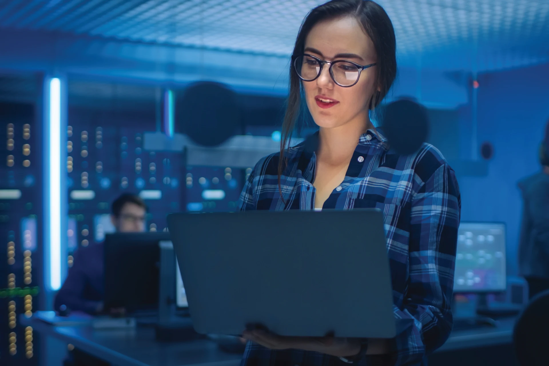 Professional holding a laptop in a tech workspace with monitors behind.