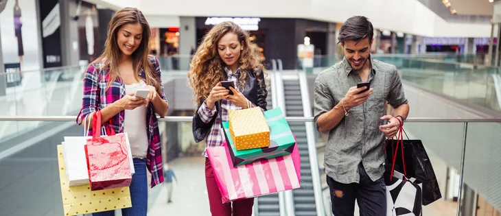 Three retail shoppers holding bags and using smartphones inside mall hallway