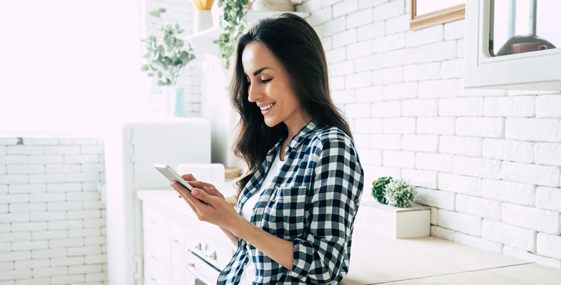 Casual woman sitting in bright kitchen space typing on smartphone screen