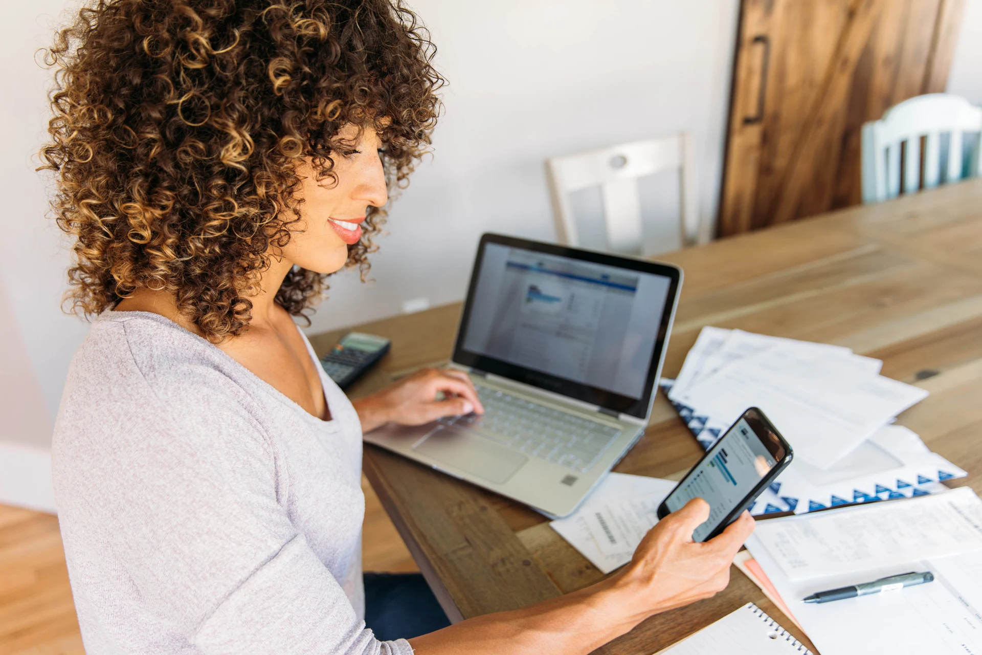 Individual checking banking app on smartphone and financial documents on laptop