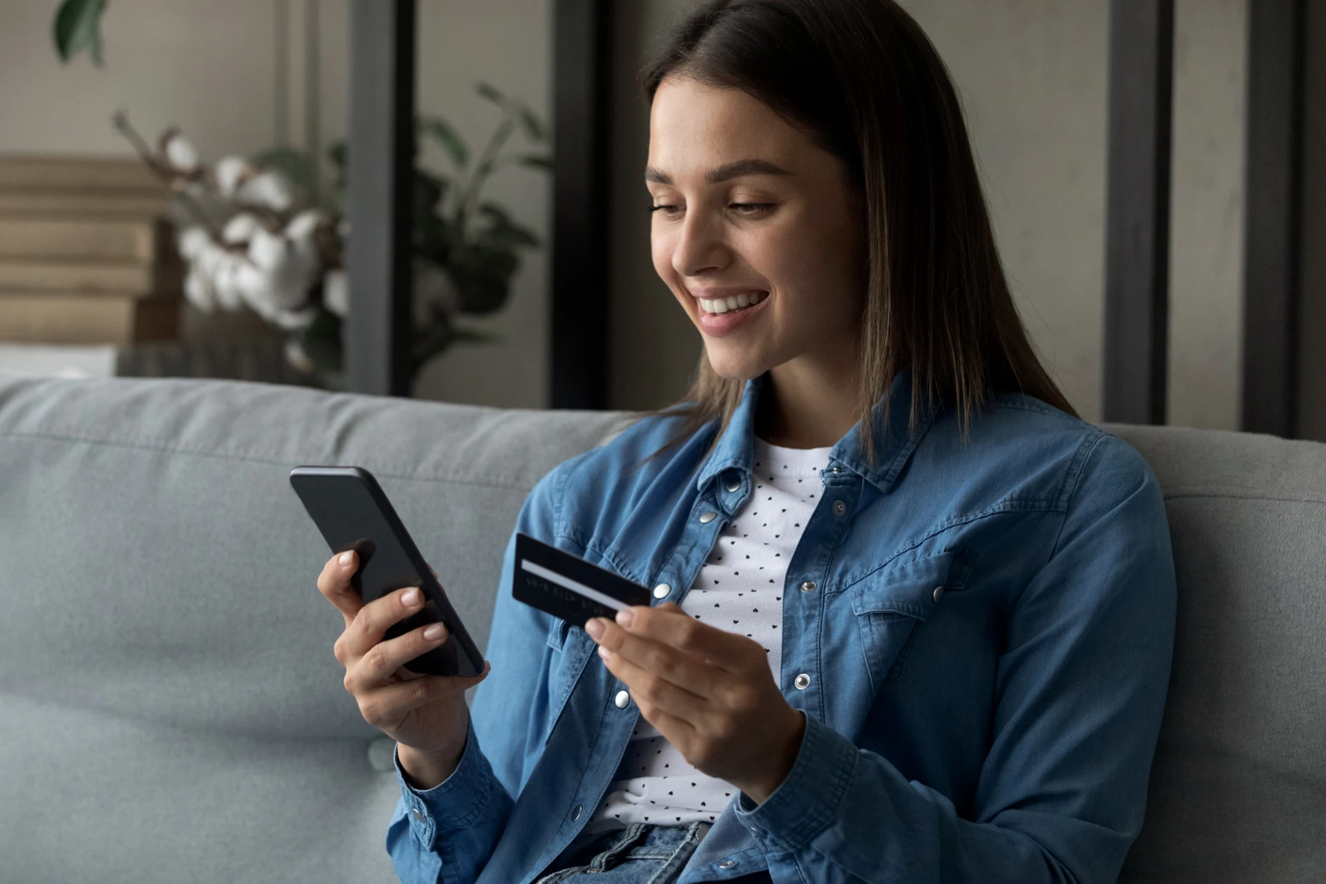 Person sitting on living room sofa using smartphone and holding credit card