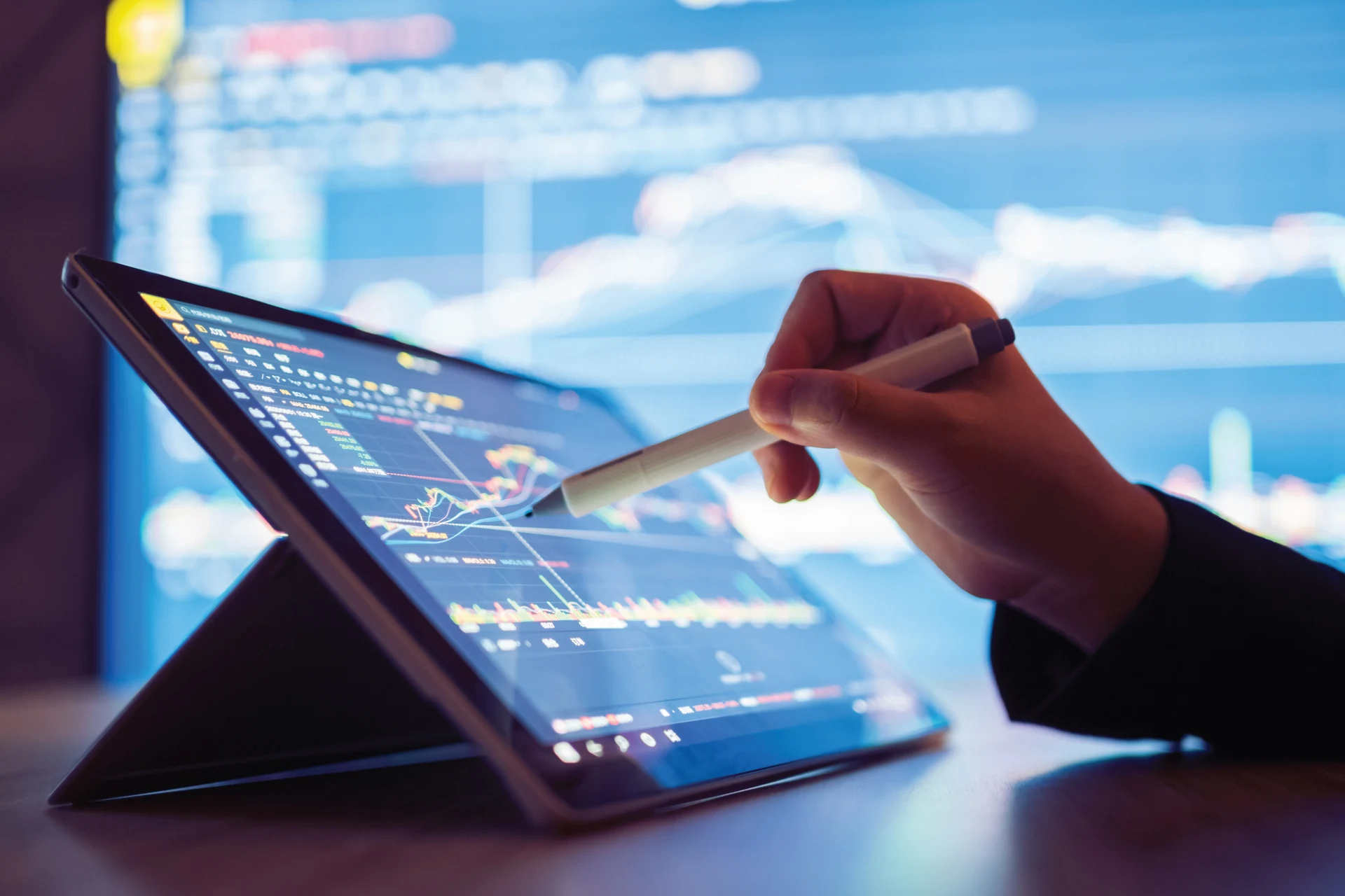 Hand using a stylus on a tablet displaying trading data analytics graphs.