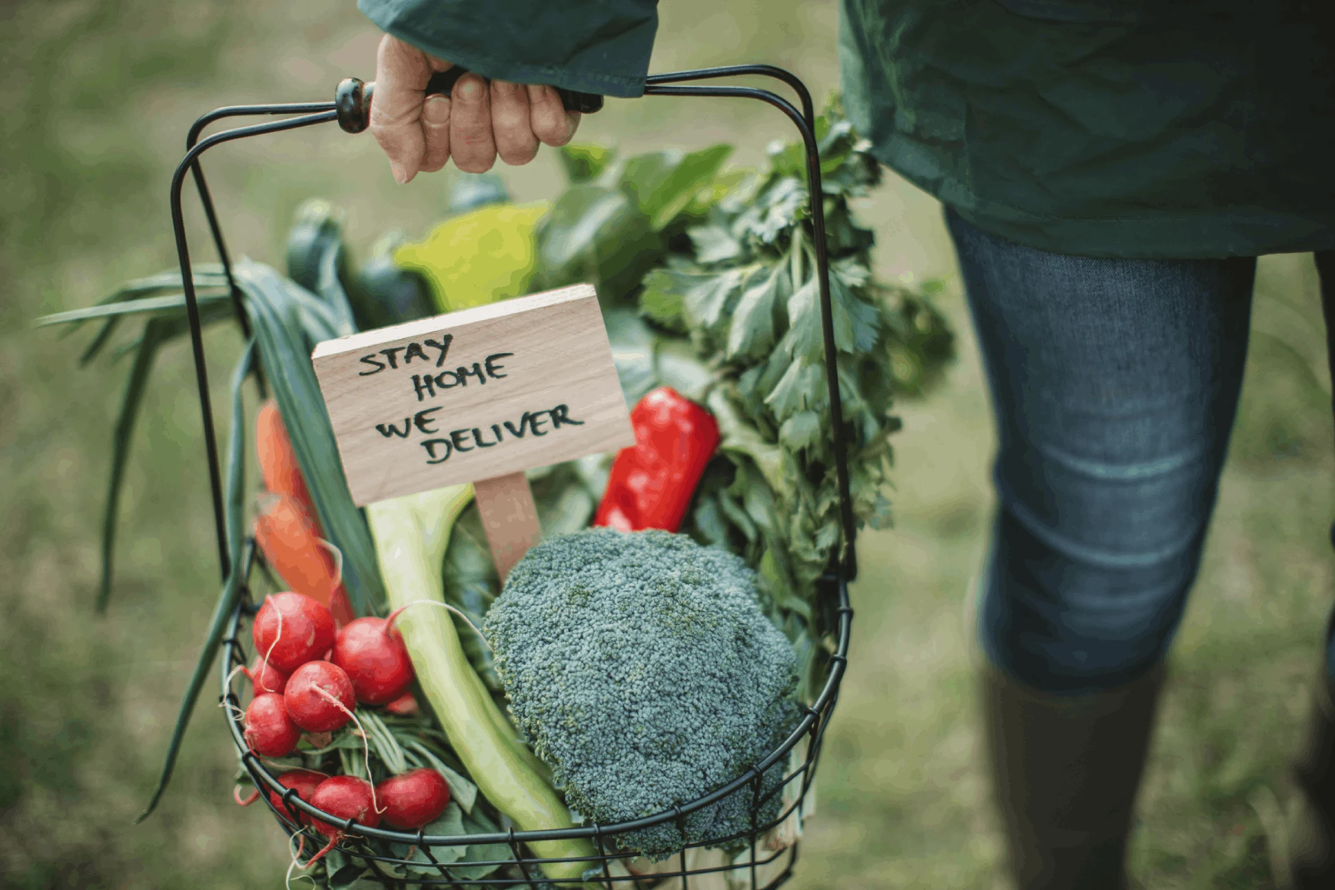 A hand holding a basket of fresh vegetables with a sign saying 'Stay Home We