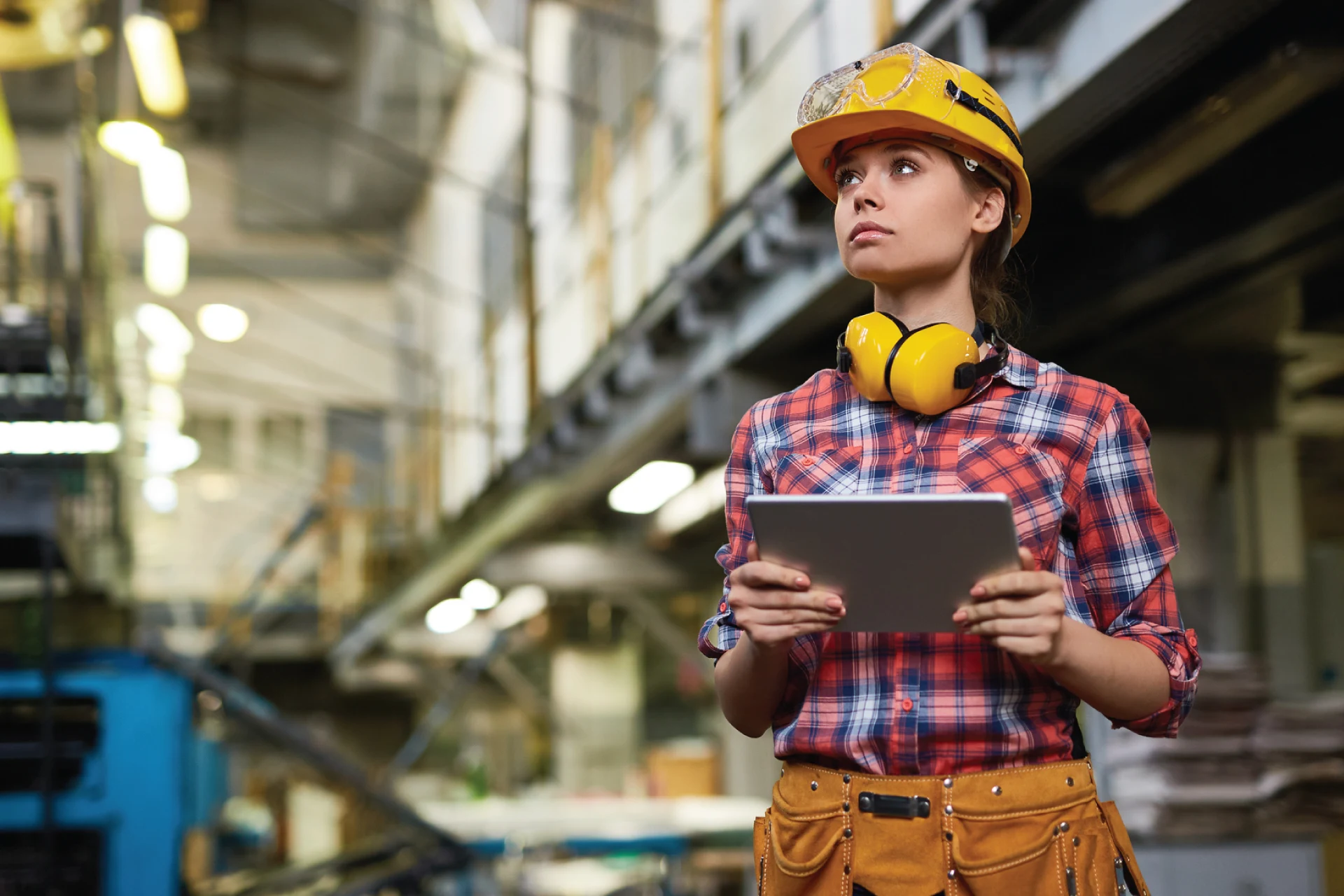 A worker in a manufacturing setting holding a tablet and wearing safety gear