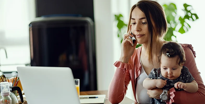 Working mother using laptop on desk while holding baby infant in arms at home