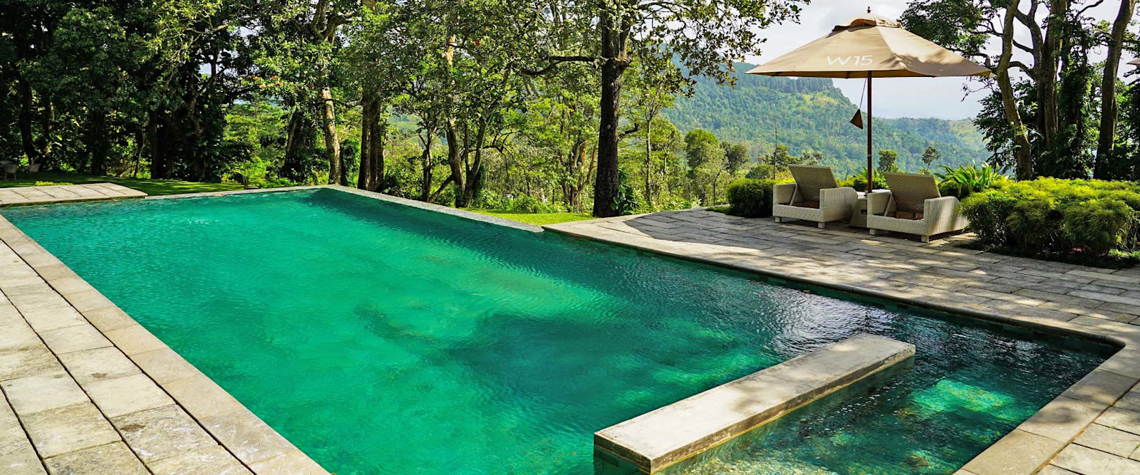 The green-tiled infinity pool on a stone terrace, with an umbrella, trees and hills beyond