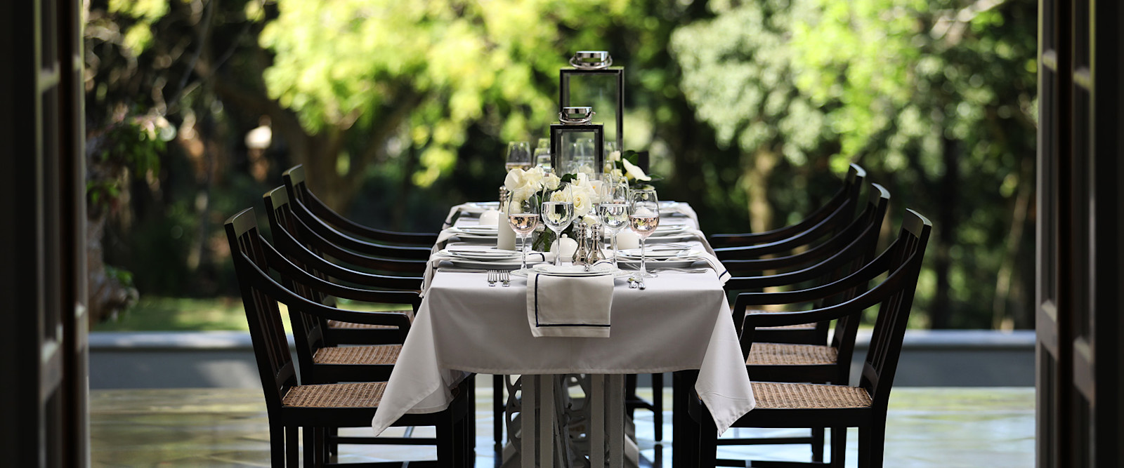 A long table laid for dinner with white linen and flowers on the terrace, facing trees