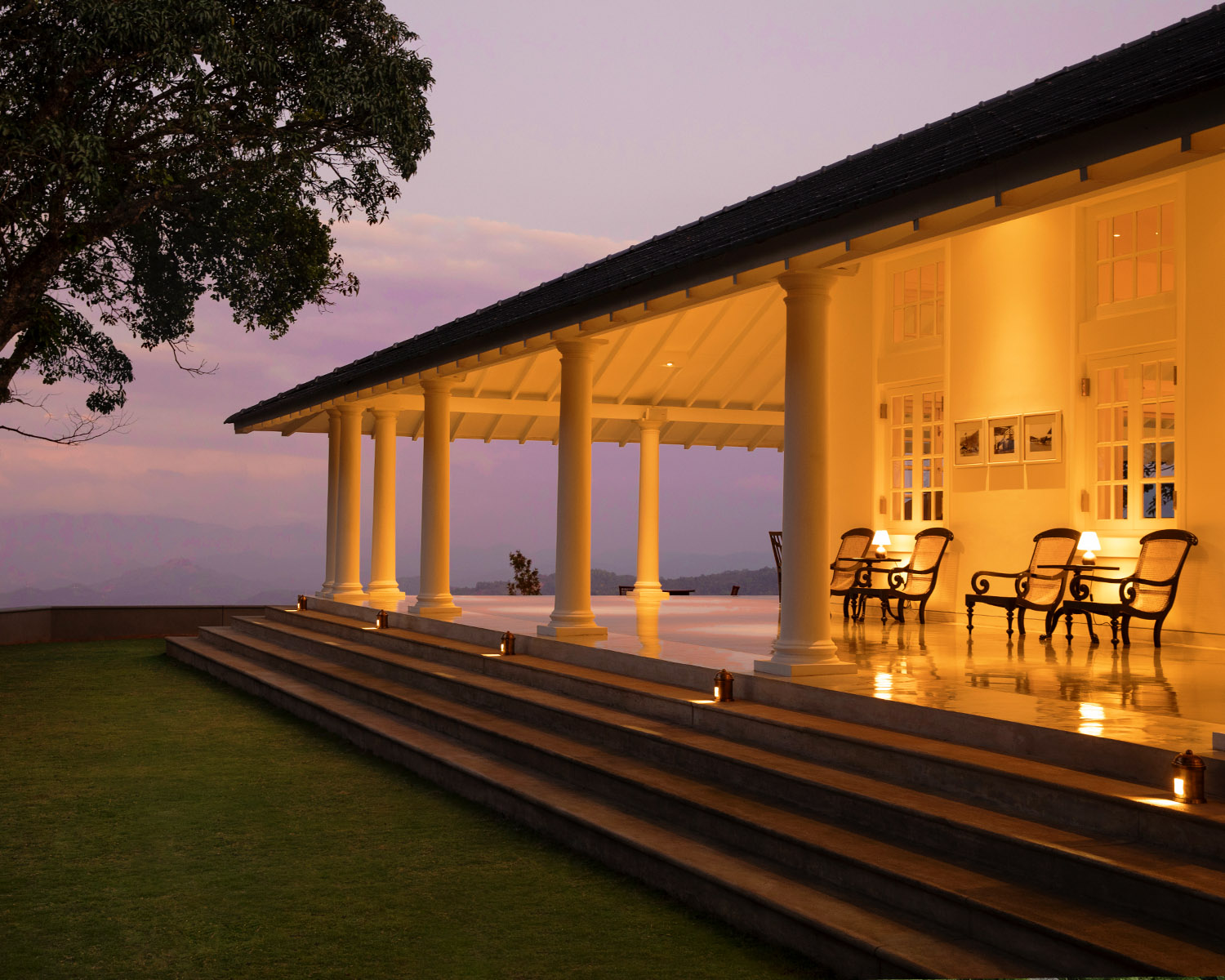 The columned verandah lit at dusk, with chairs, steps down to the lawn and a large tree