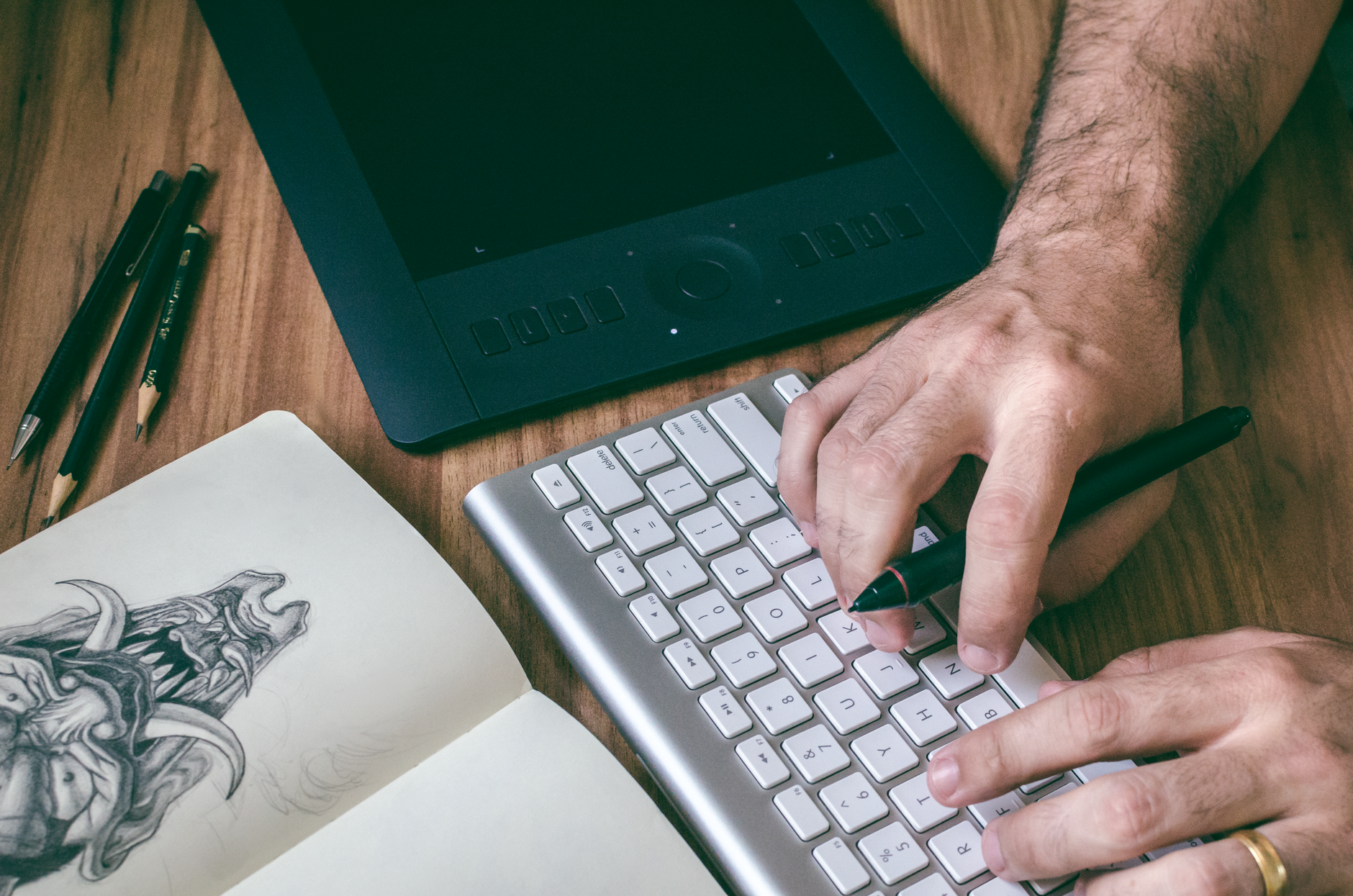 A designer's hands working on digital art with a tablet, keyboard, and sketchbook.