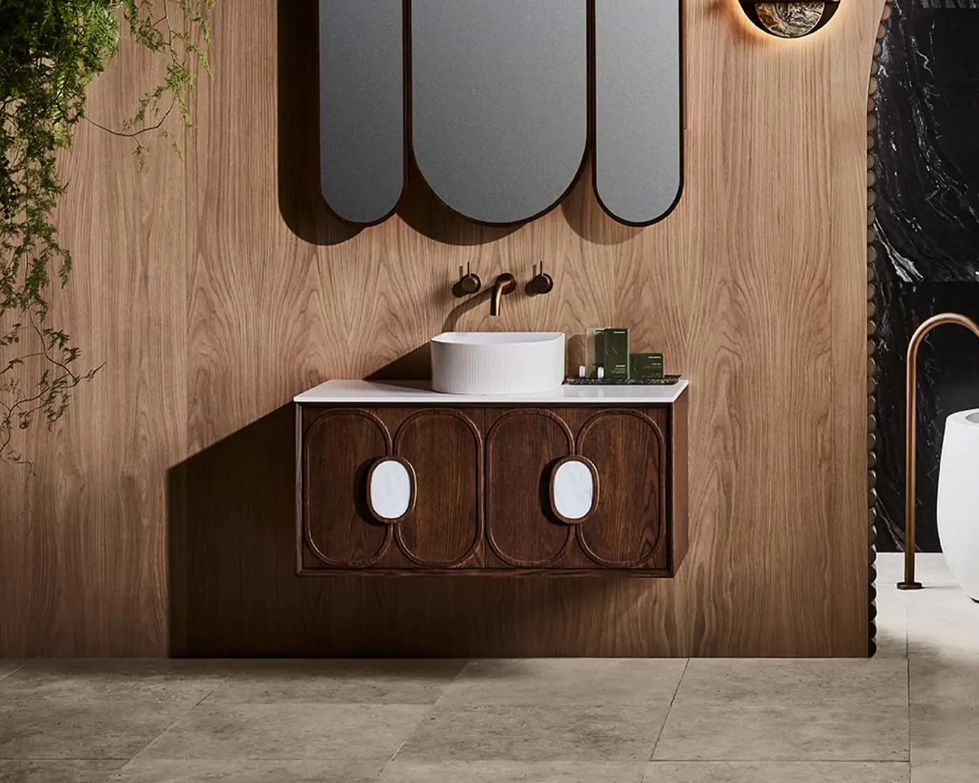 Modern bathroom with wooden vanity, white vessel sink, bronze fixtures, oval mirrors, and plants against wood-paneled wall.