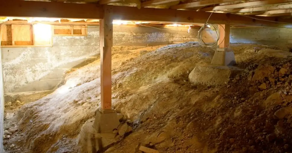 Unfinished crawl space with dirt floor, wooden support beams, and exposed foundation walls lit by utility lights.