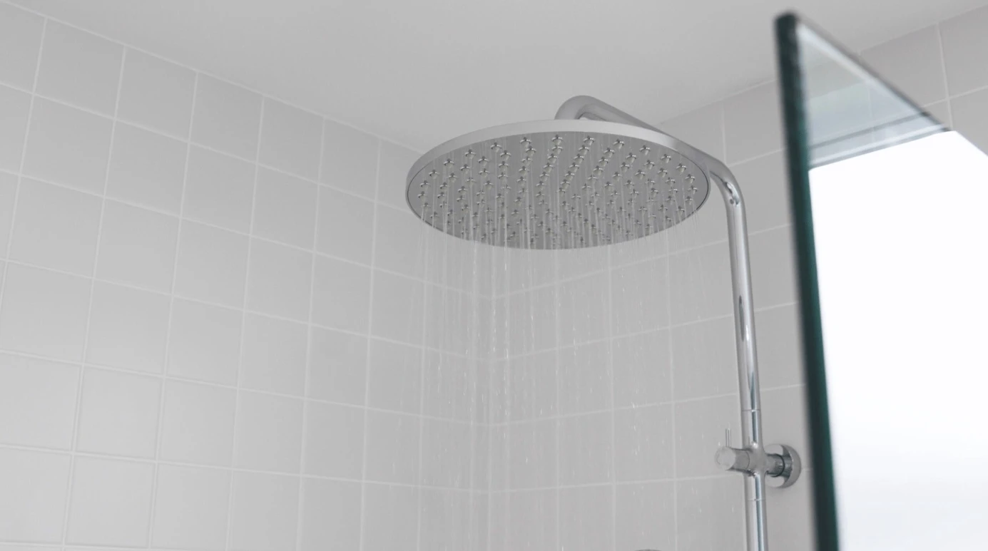 Large circular rainfall showerhead mounted on chrome pipe in white tiled bathroom with water streaming down.