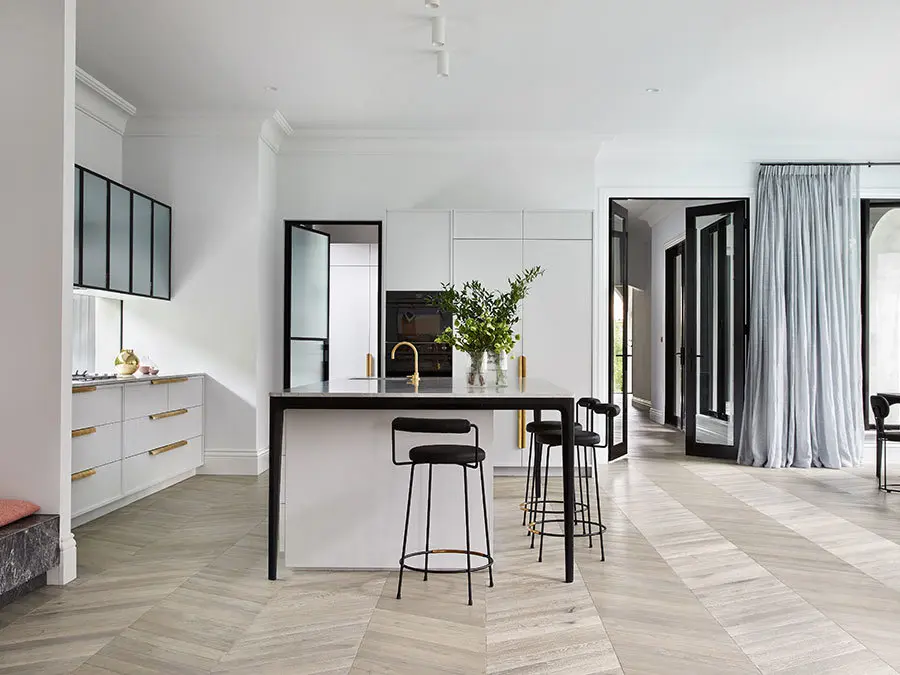 Modern white kitchen with black island, gold fixtures, black barstools, herringbone wood floors, and light blue curtains.