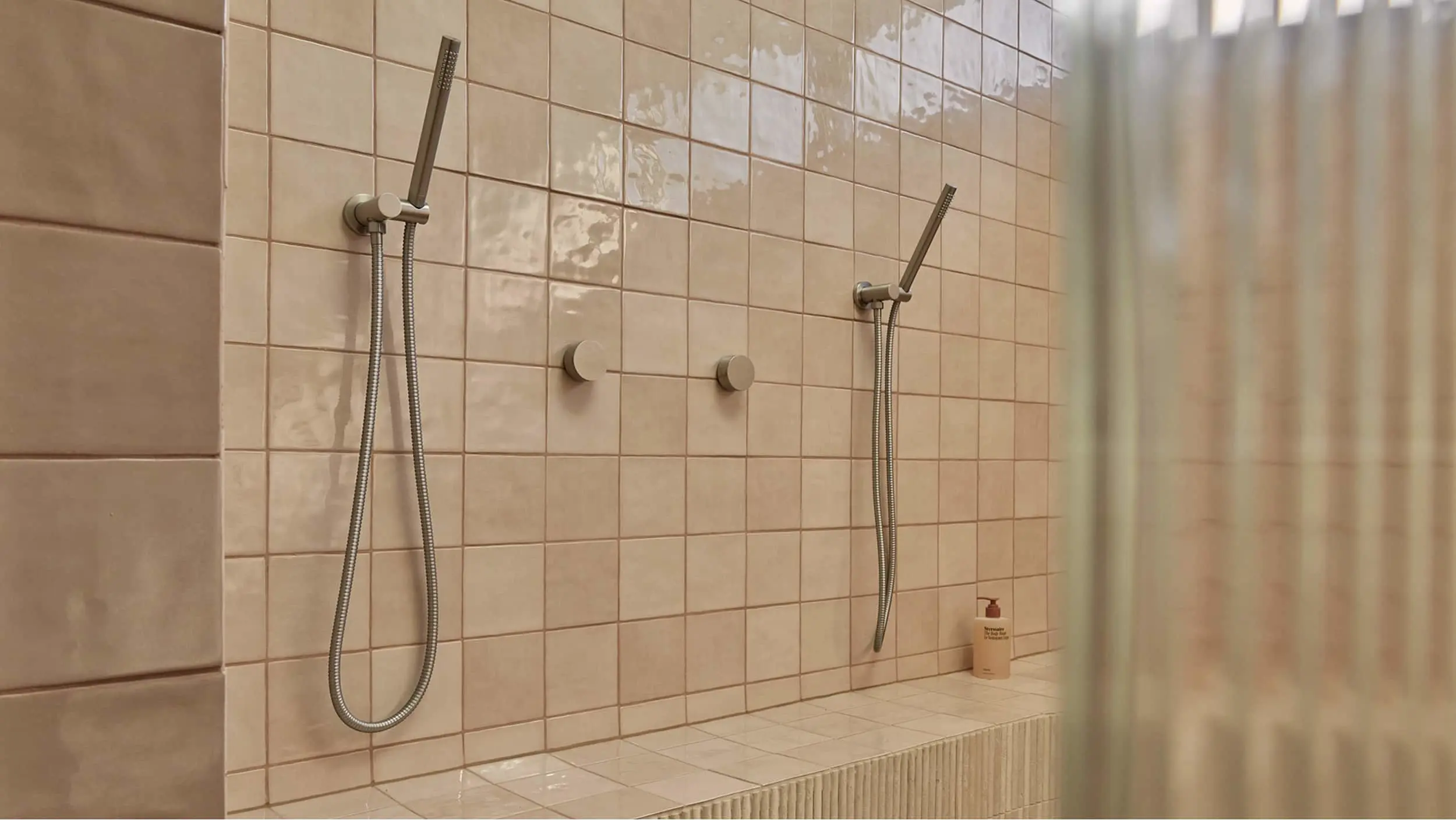 Modern shower with beige tiled walls featuring two handheld showerheads, control knobs, and a small bottle of product.