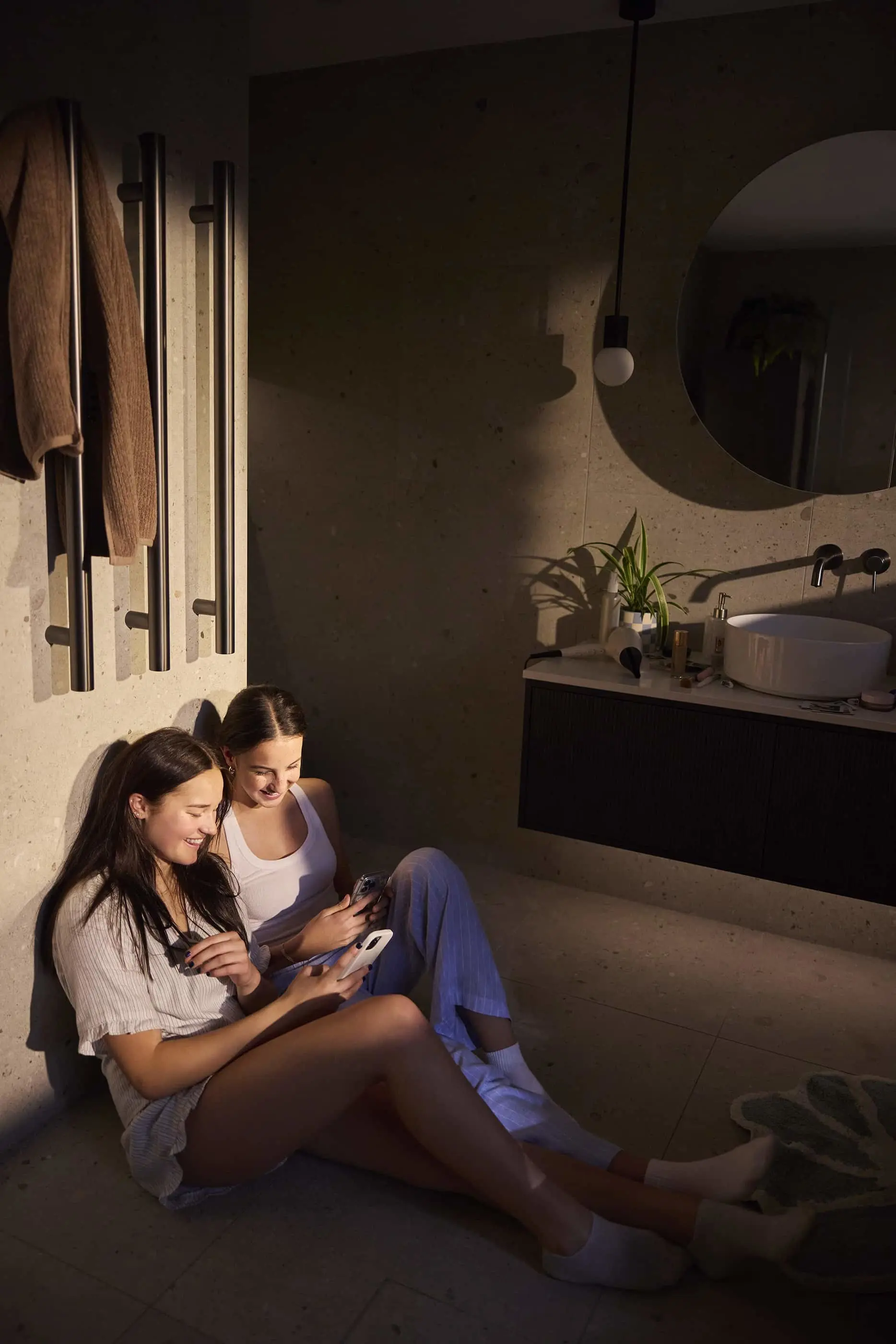 Two people sitting on bathroom floor looking at phones, with modern sink, round mirror, and towel rack visible.