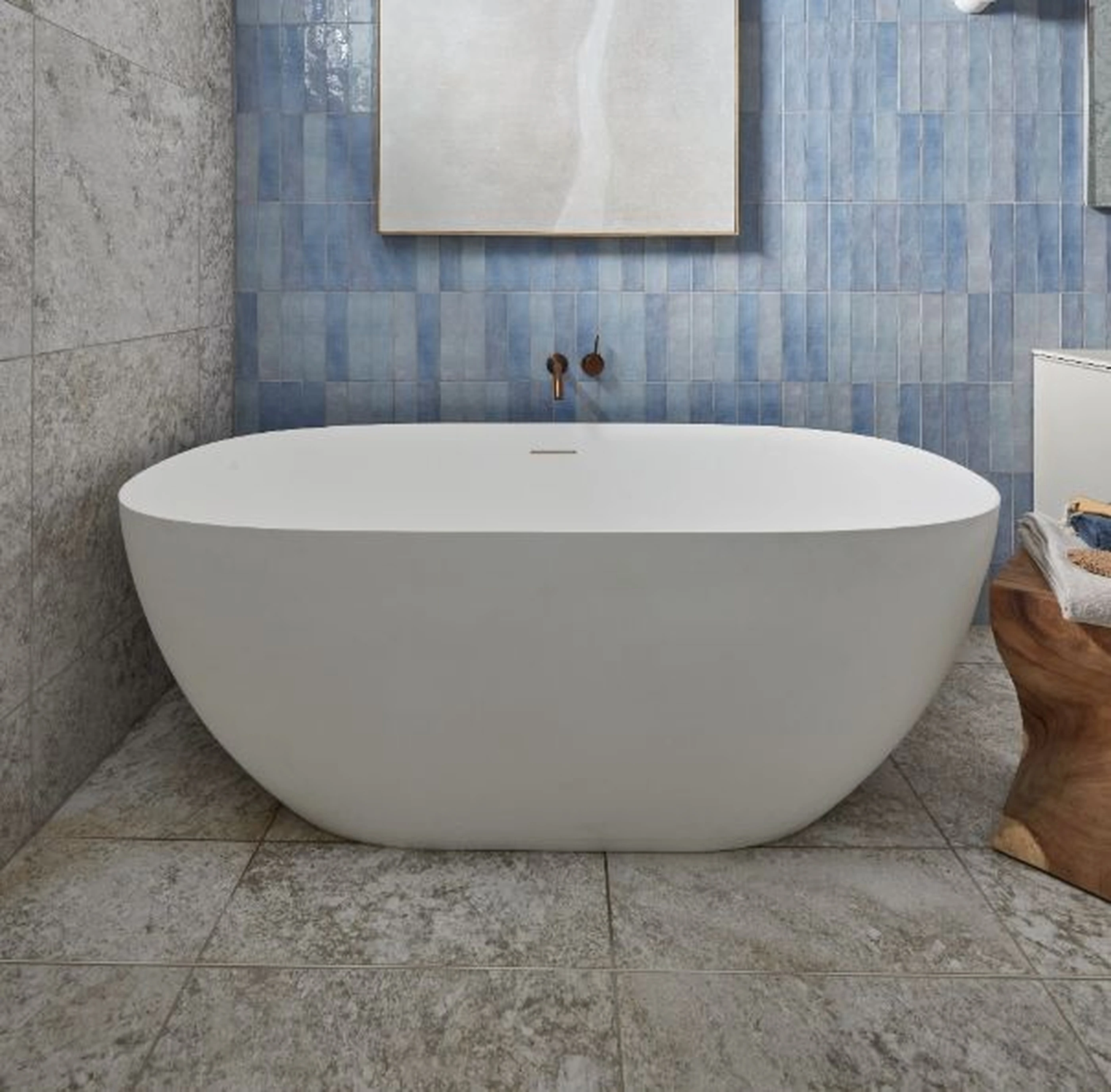 Modern white freestanding bathtub against blue tile wall with framed artwork and brass wall-mounted faucet.