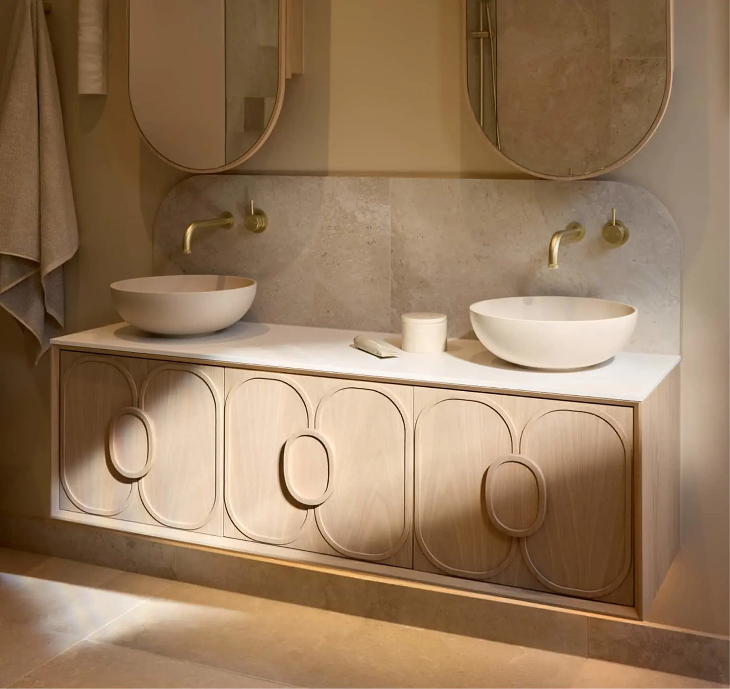 Floating wood bathroom vanity with oval door panels, two white vessel sinks, brass wall-mount faucets, and two rounded mirrors above.