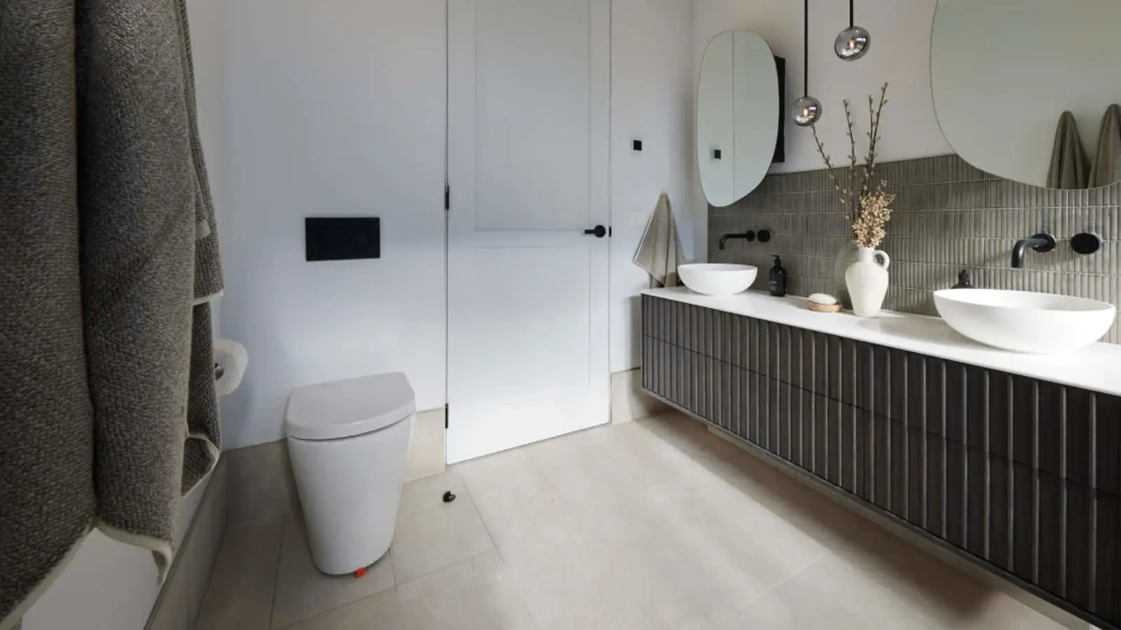 Modern bathroom with white toilet, gray vanity with vessel sinks, oval mirrors, and pendant lighting.