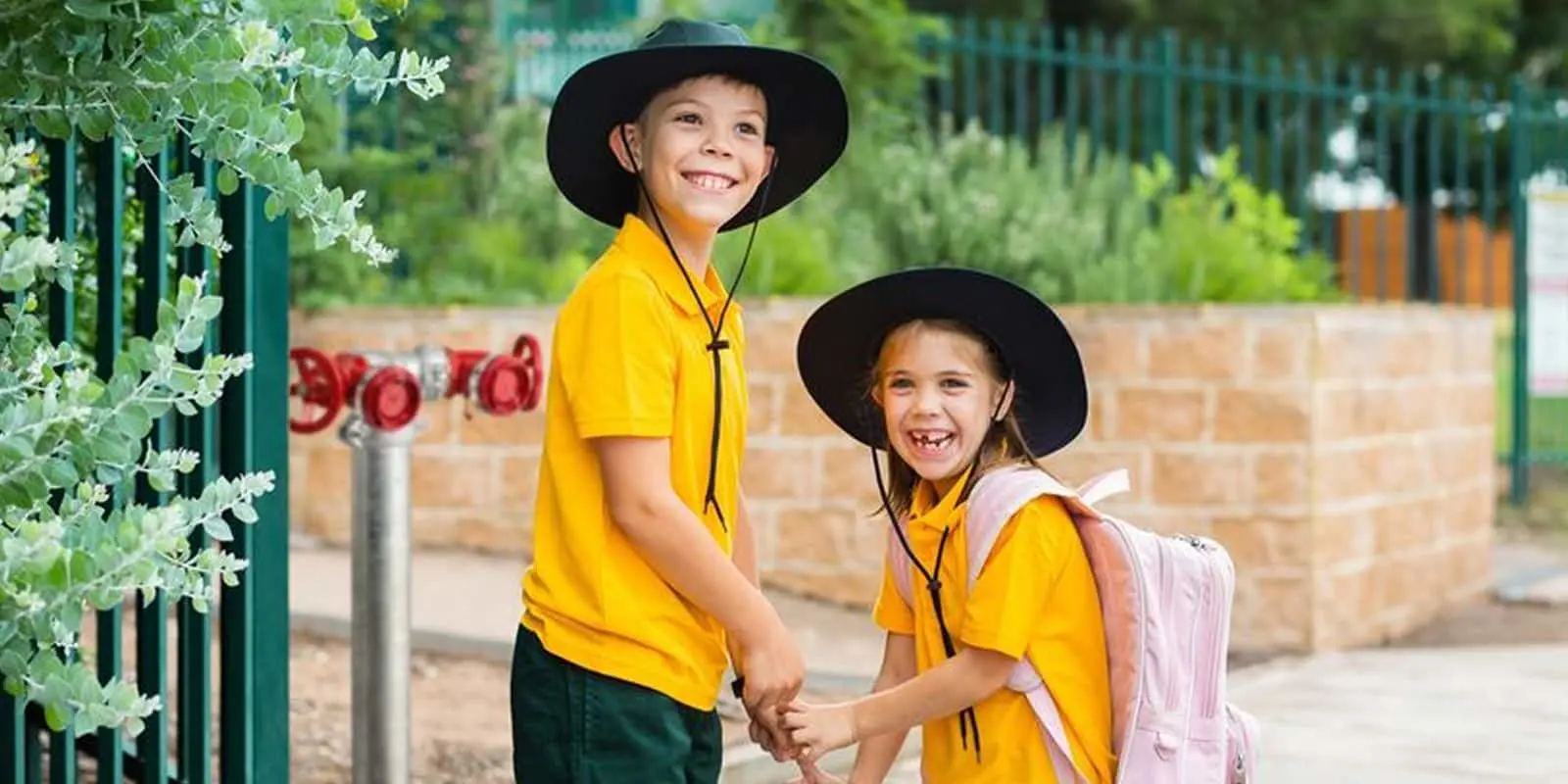 Two smiling children in yellow school uniforms and wide-brimmed black hats holding hands outside a school gate.