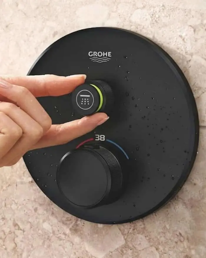 A hand pressing a button on a round matte black GROHE shower thermostat with two knobs, mounted on a stone wall.